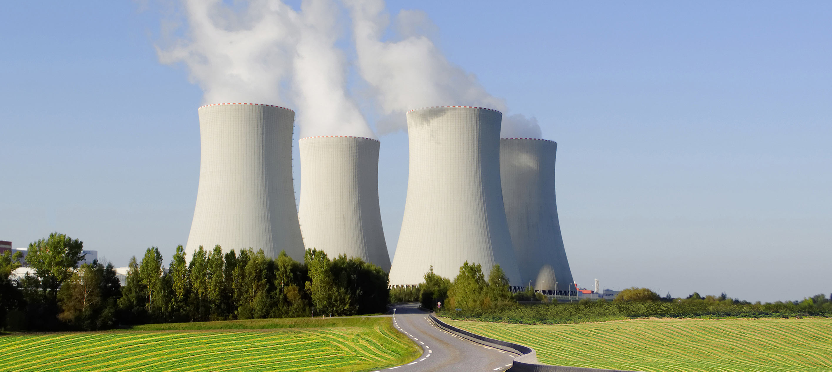 Cooling towers of a nuclear power plant emitting steam, set against a clear blue sky with green fields and a curved road in front.
