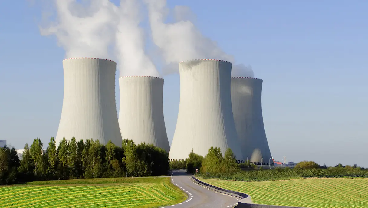 Cooling towers of a nuclear power plant emitting steam, set against a clear blue sky with green fields and a curved road in front.