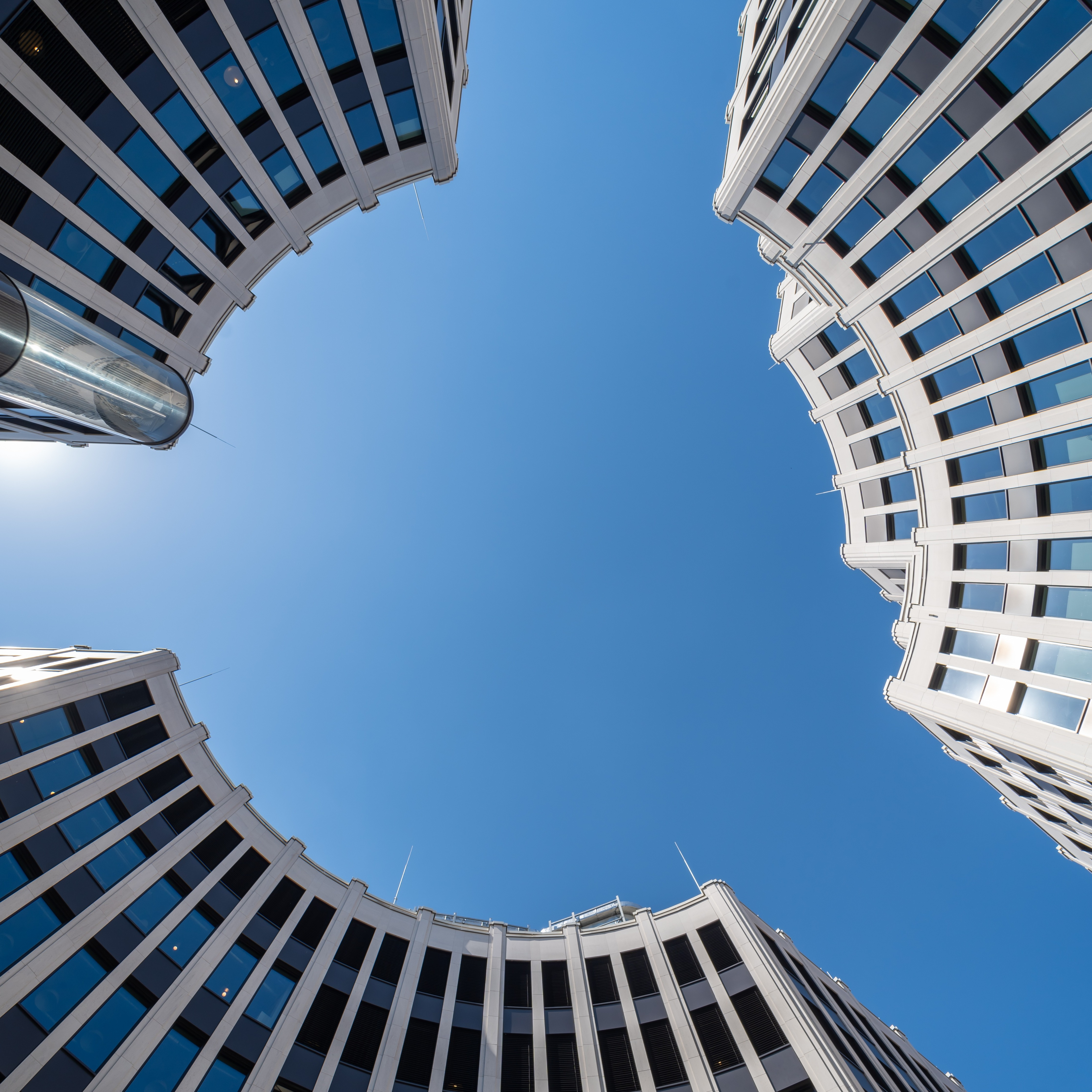 Curved modern office buildings with white facades and dark windows frame a clear blue sky, viewed from below.