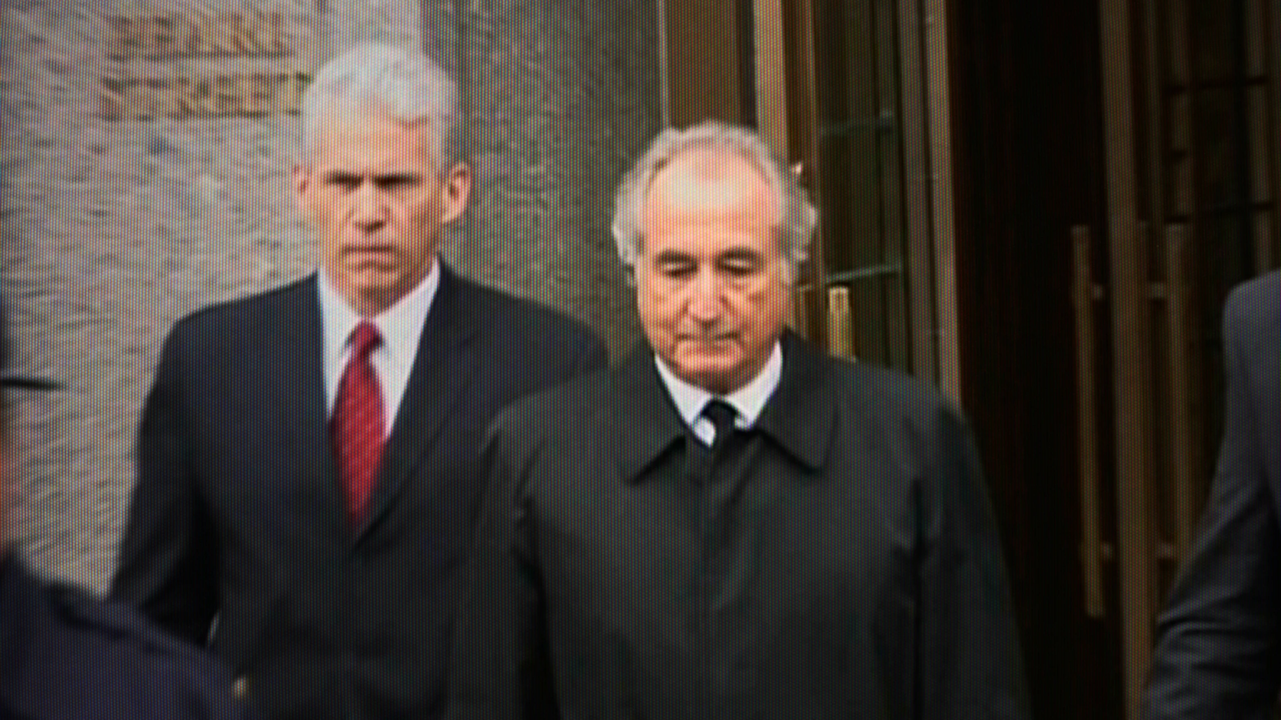 Two older men in formal suits and overcoats are walking outside a building with glass and stone walls.