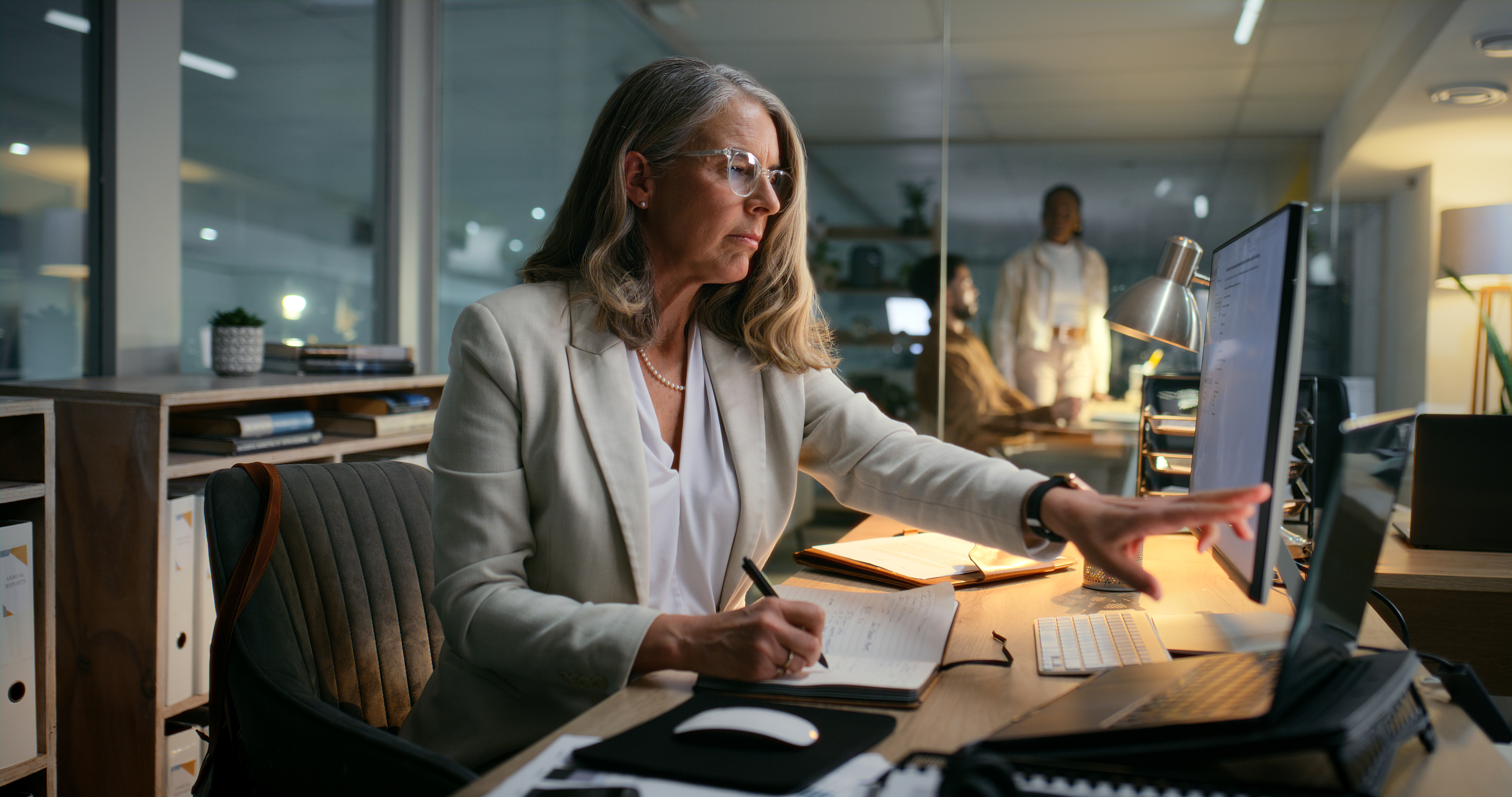 A woman in a white blazer is working at a desk with a notebook, pen, and computer in a modern office during the evening with two colleagues in the background.