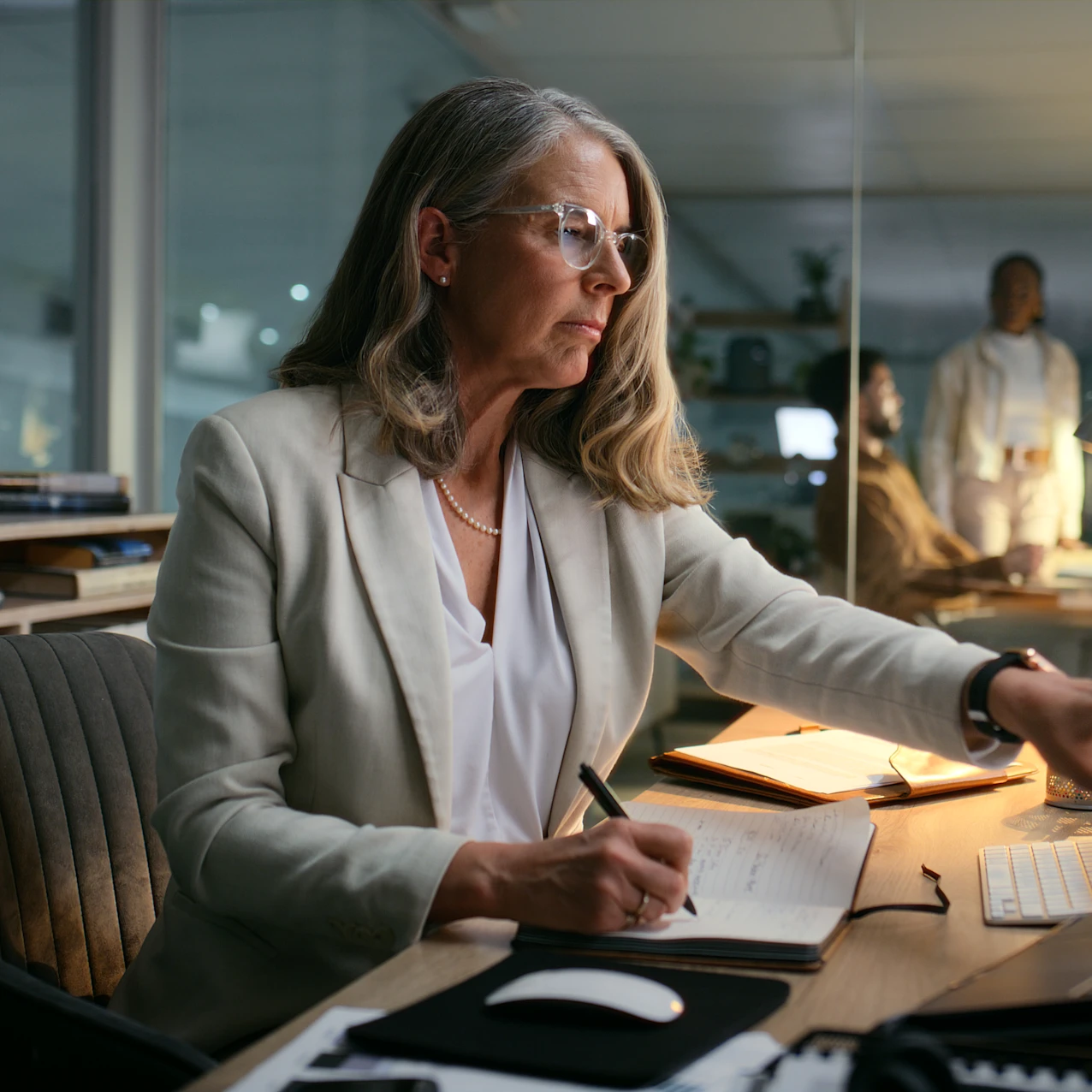 A woman in a white blazer is working at a desk with a notebook, pen, and computer in a modern office during the evening with two colleagues in the background.