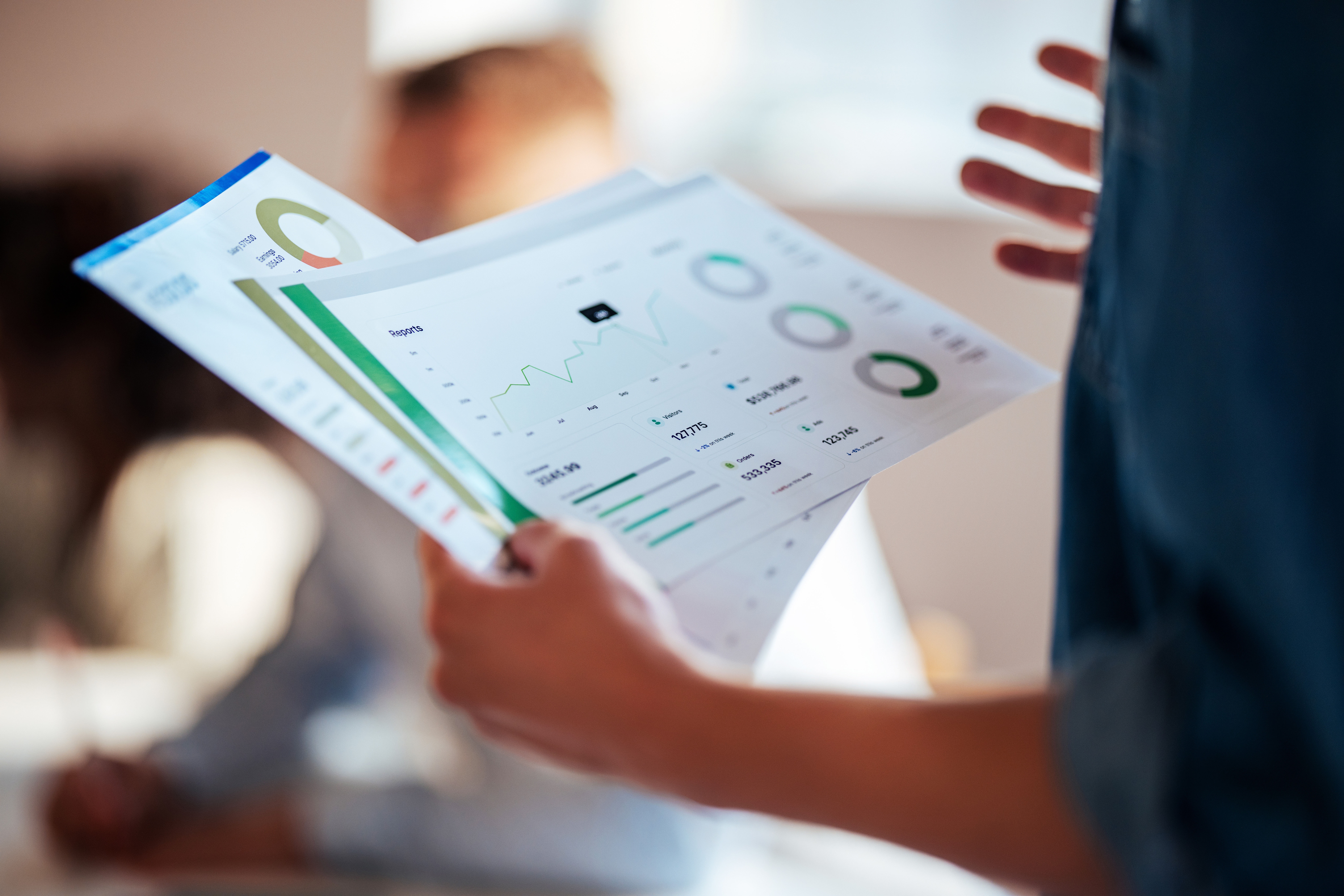 Person holding and reviewing colorful business charts and graphs printed on paper with blurred figures in the background.