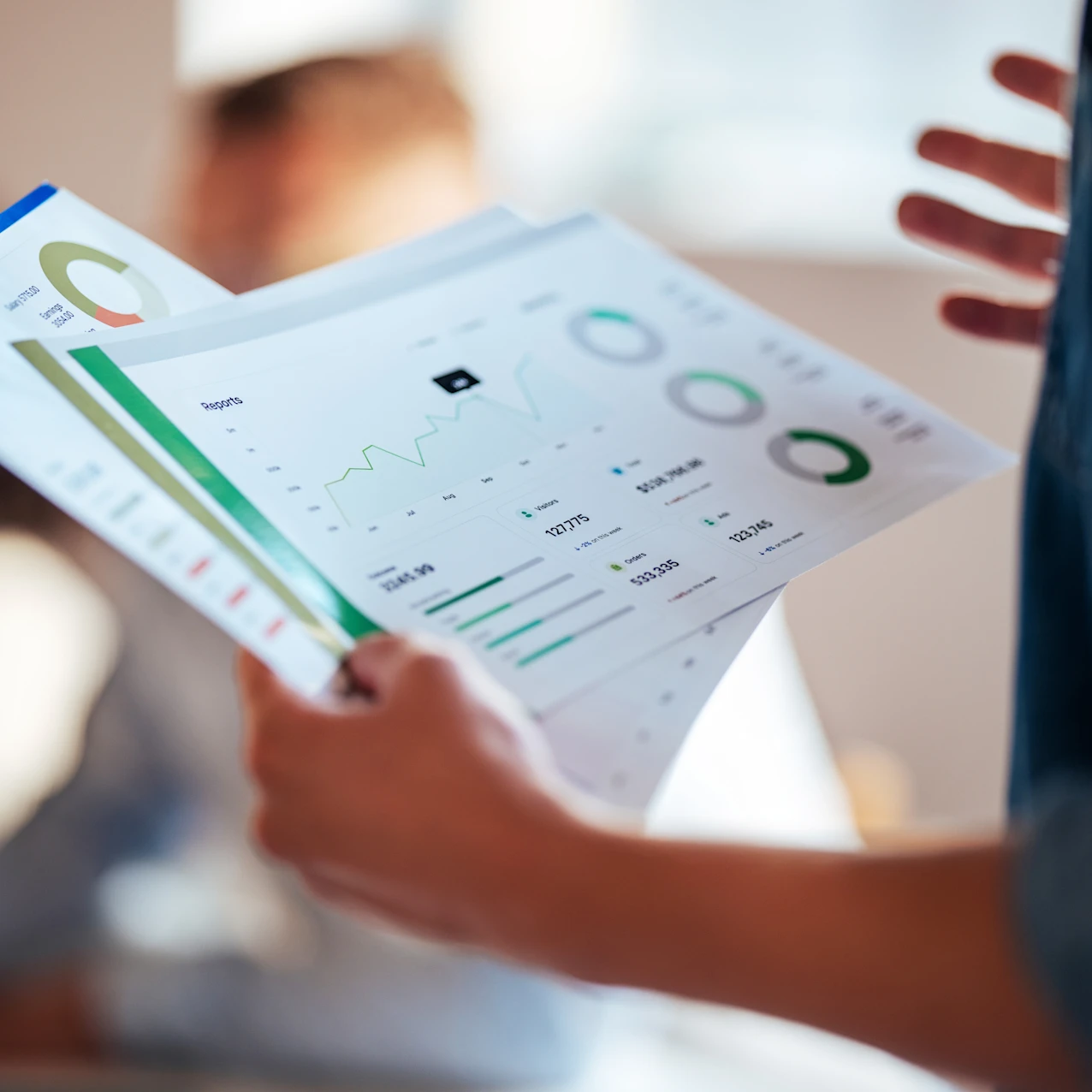Person holding and reviewing colorful business charts and graphs printed on paper with blurred figures in the background.