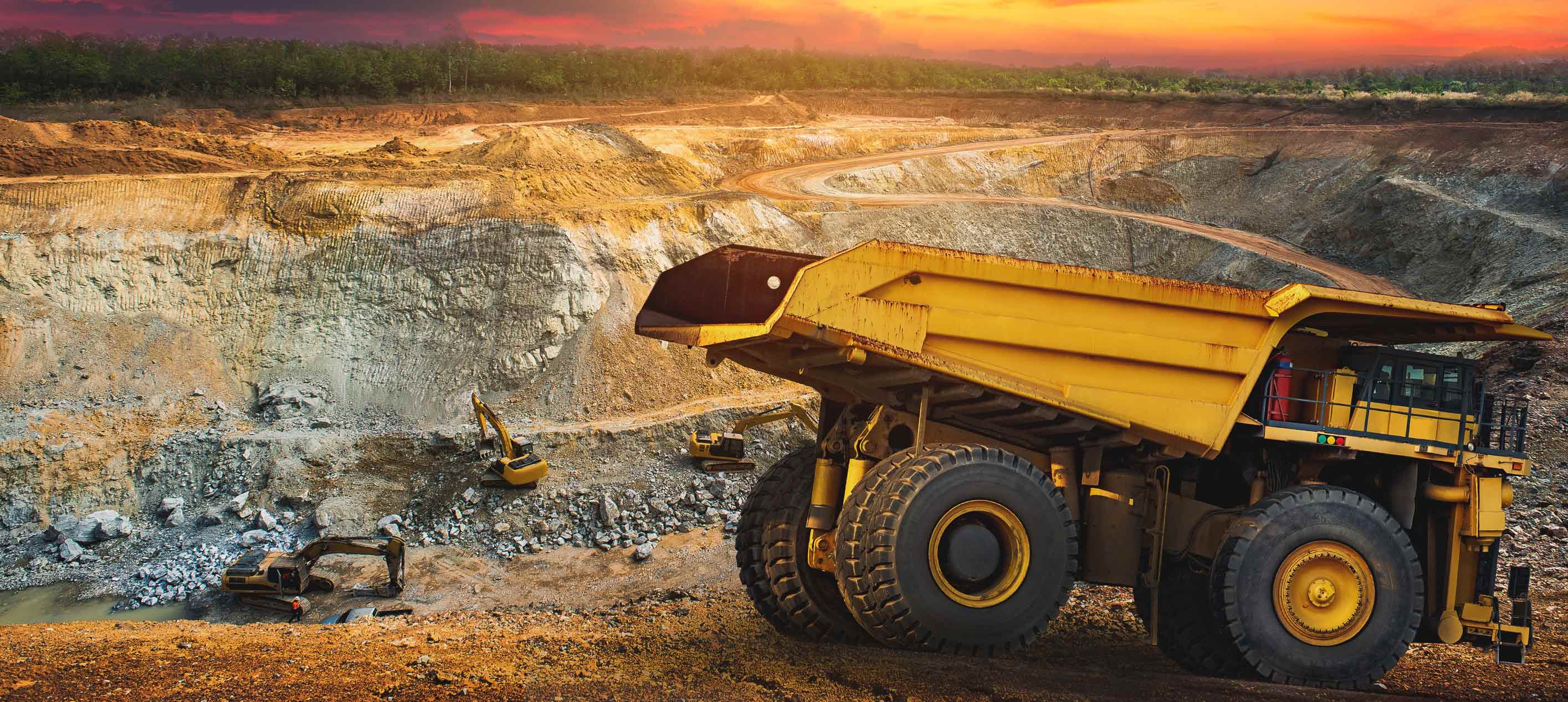 Large yellow dump truck and excavators operating in an open-pit mining site with winding roads and a sunset sky in the background.