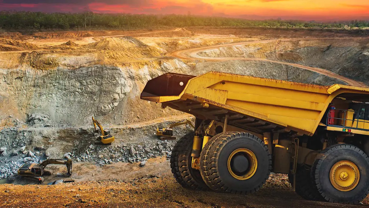 Large yellow dump truck and excavators operating in an open-pit mining site with winding roads and a sunset sky in the background.