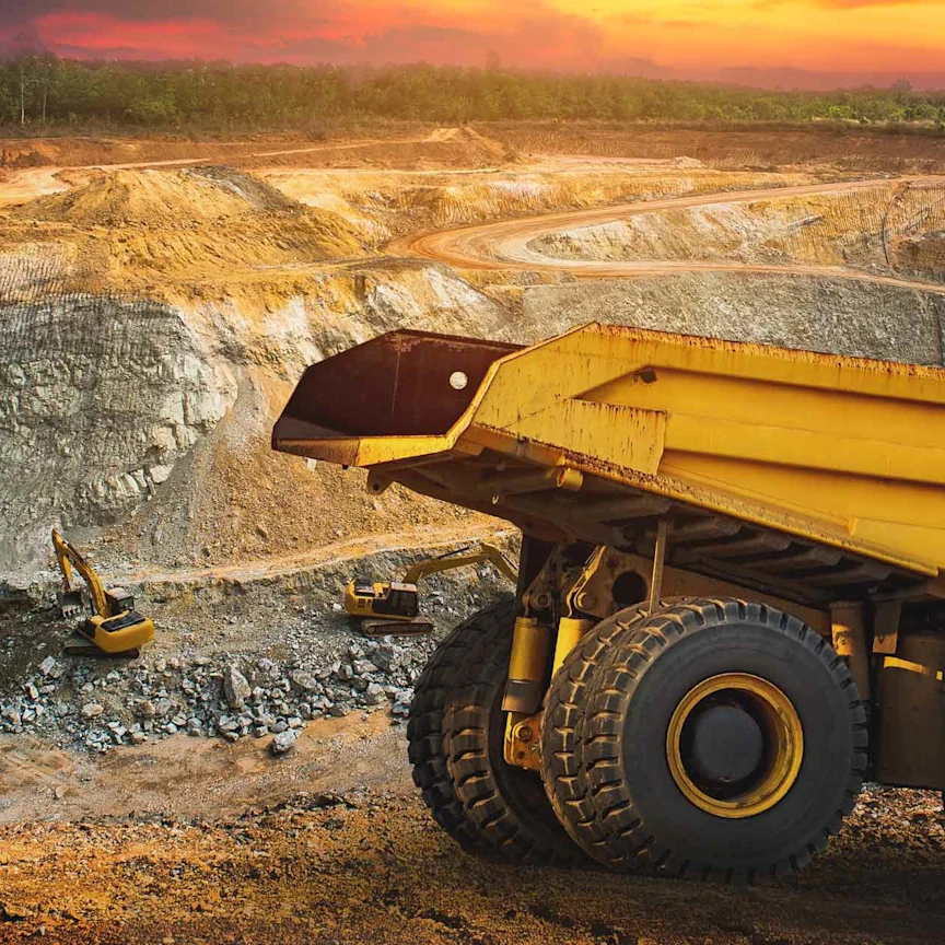 Large yellow dump truck and excavators operating in an open-pit mining site with winding roads and a sunset sky in the background.