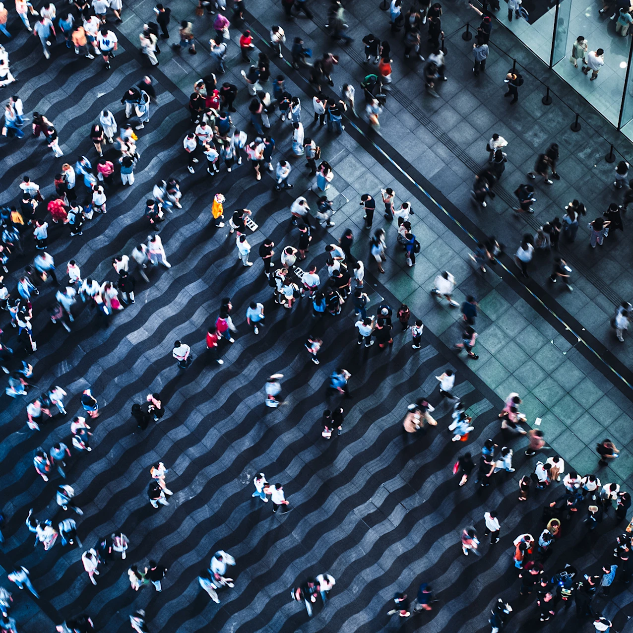 Aerial view of a crowded urban plaza with people walking on a dark wavy-patterned pavement near a modern glass building.