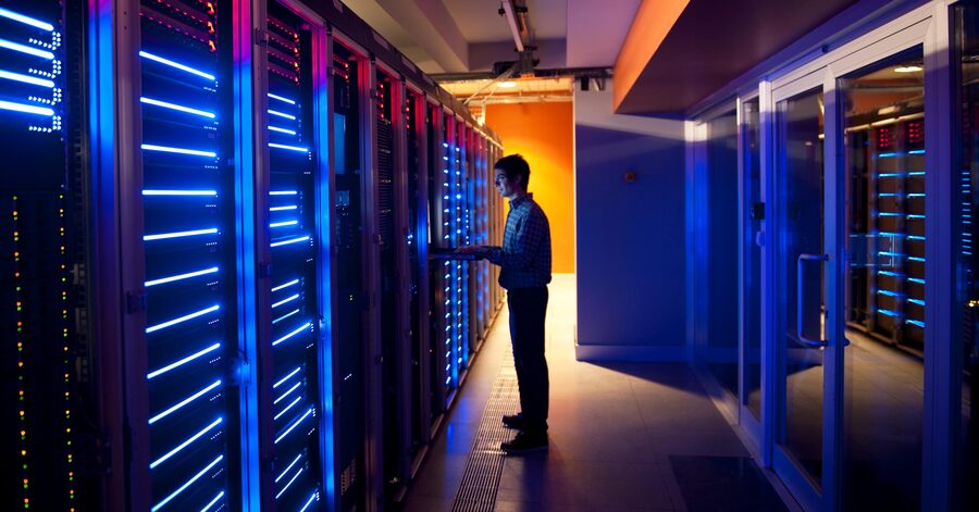 A man working on a laptop in a data centre, standing beside rows of illuminated server racks with blue and red lights.