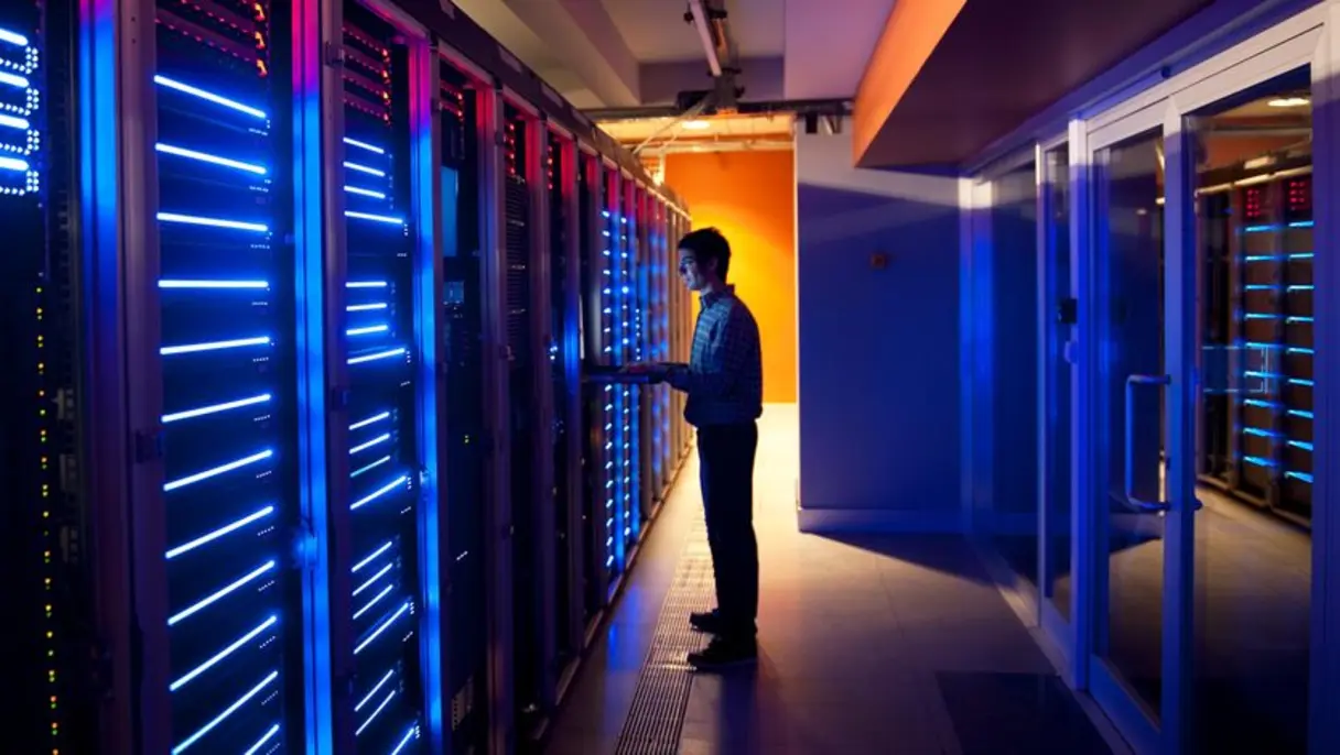 A man working on a laptop in a data centre, standing beside rows of illuminated server racks with blue and red lights.
