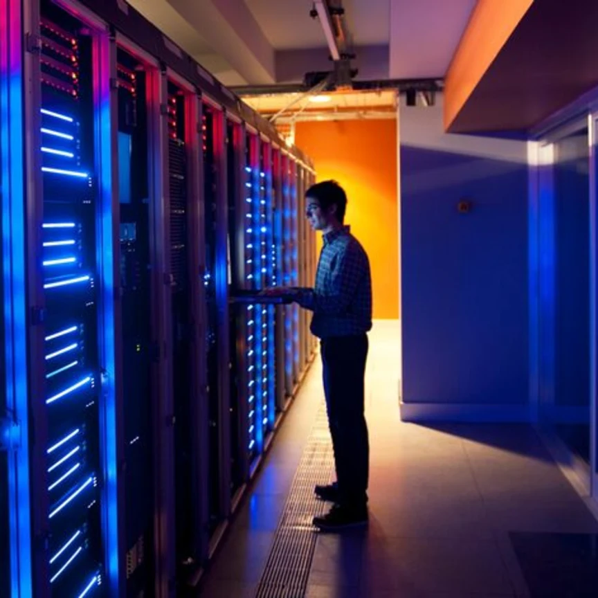 A man working on a laptop in a data centre, standing beside rows of illuminated server racks with blue and red lights.