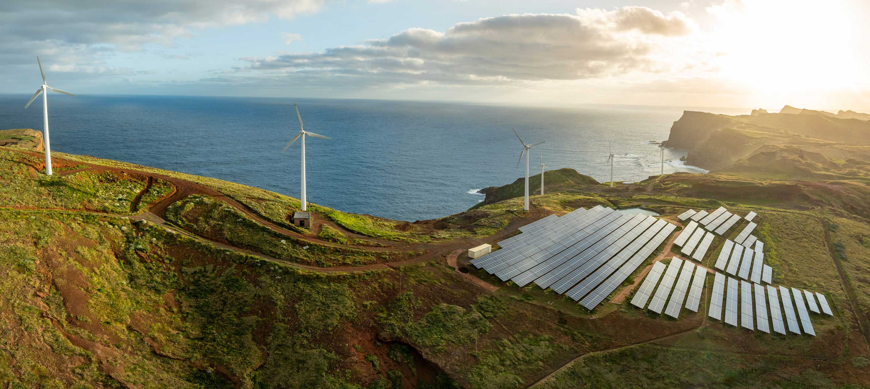 Wind turbines and solar panel arrays on coastal hills overlooking the ocean at sunset with dirt access roads.