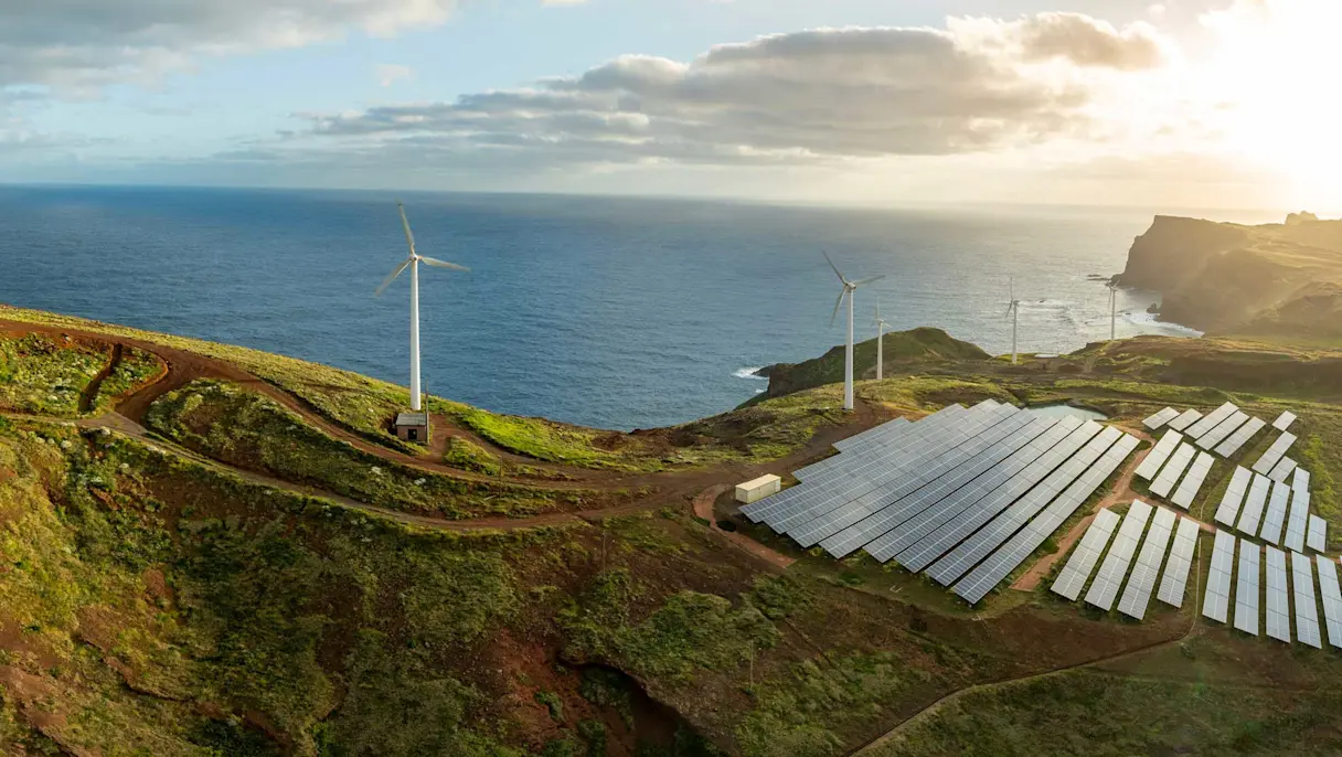 Wind turbines and solar panel arrays on coastal hills overlooking the ocean at sunset with dirt access roads.