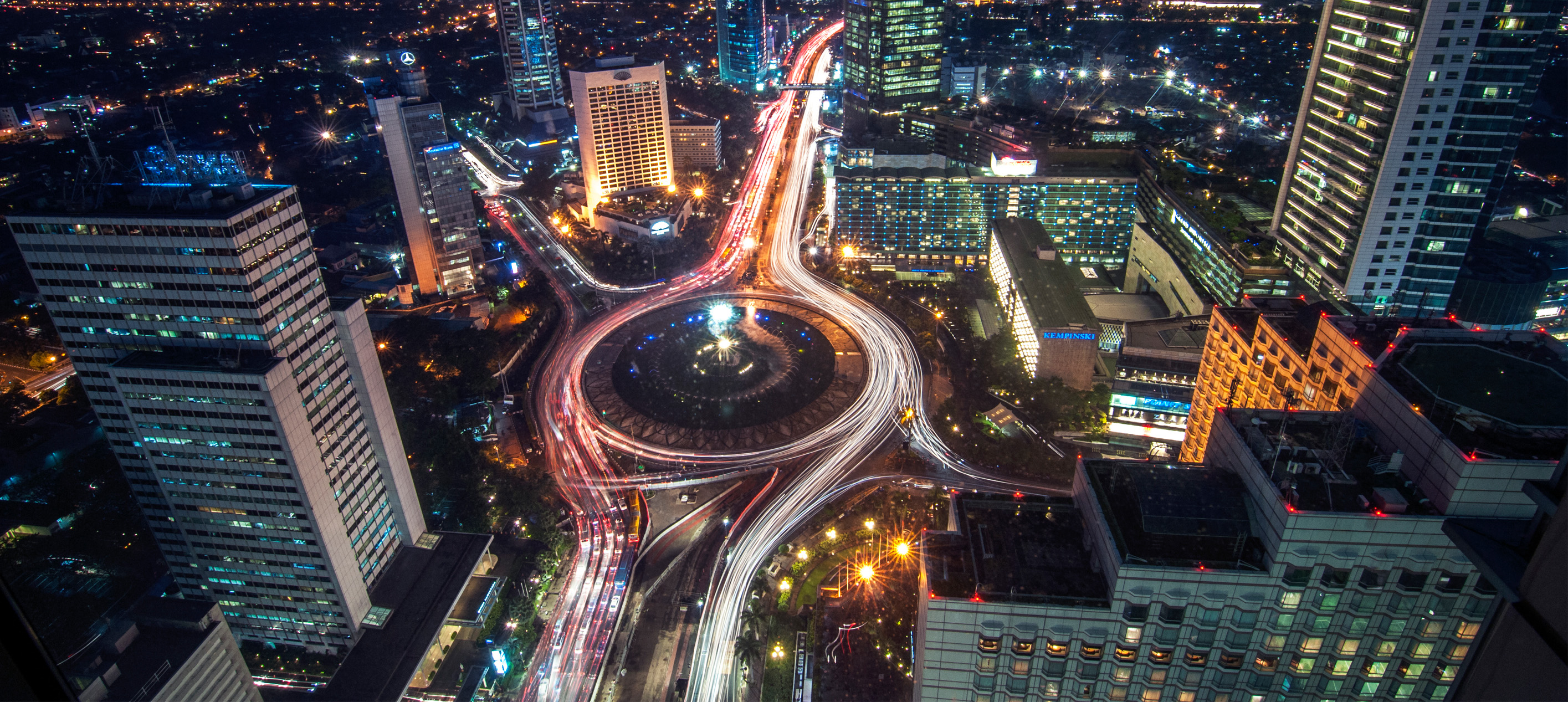Nighttime aerial view of a modern city skyline with illuminated skyscrapers, a large roundabout and long-exposure traffic light trails winding through the urban center.