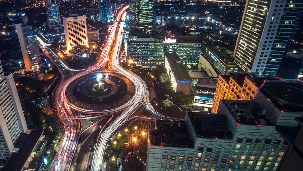 Nighttime aerial view of a modern city skyline with illuminated skyscrapers, a large roundabout and long-exposure traffic light trails winding through the urban center.