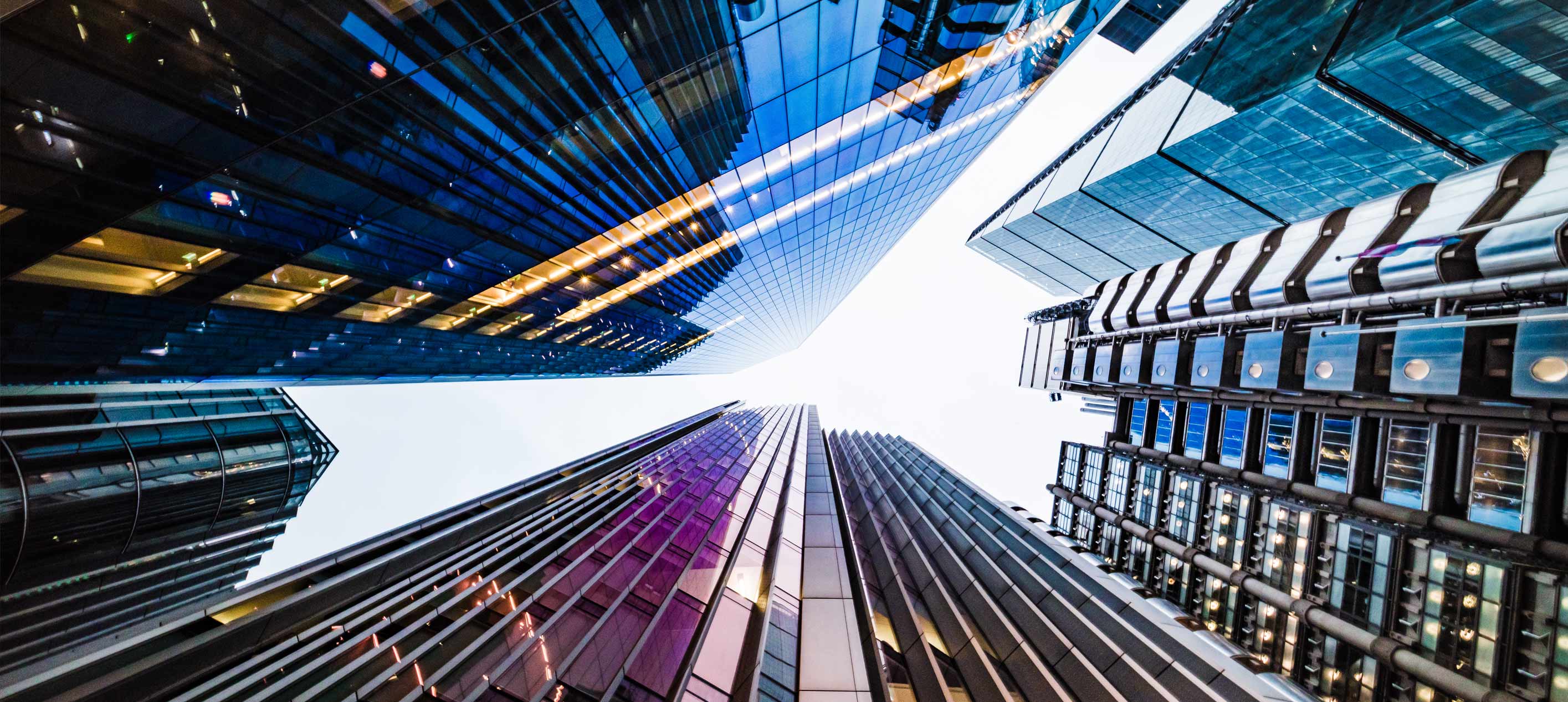 Upward view of glass skyscrapers with reflective facades against a bright sky, seen from street level below.