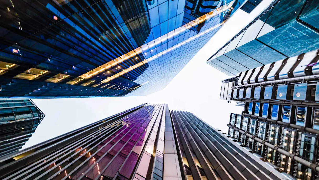 Upward view of glass skyscrapers with reflective facades against a bright sky, seen from street level below.