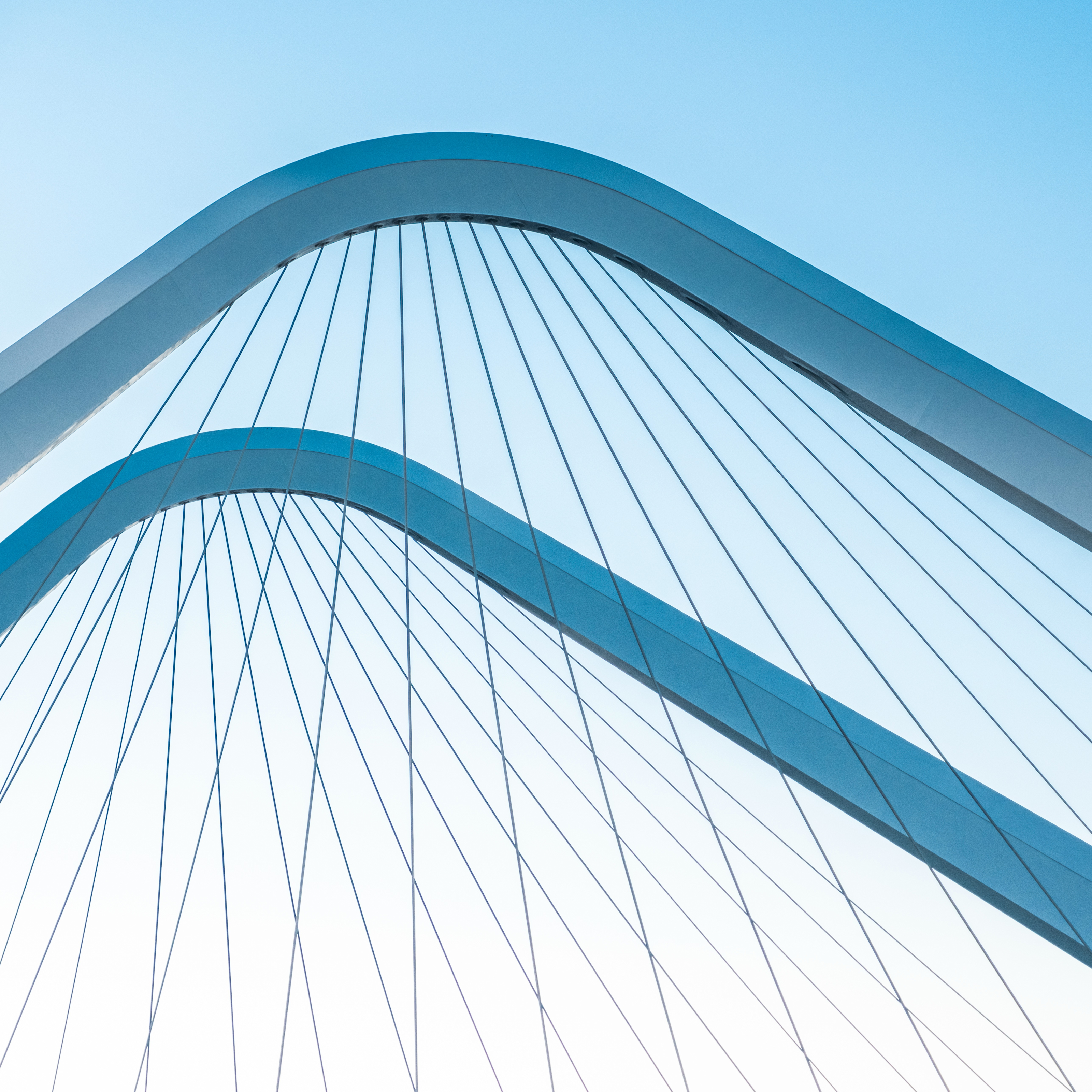 Close-up of two curved white metal arches with parallel cables against a clear blue sky, forming a modern bridge design.