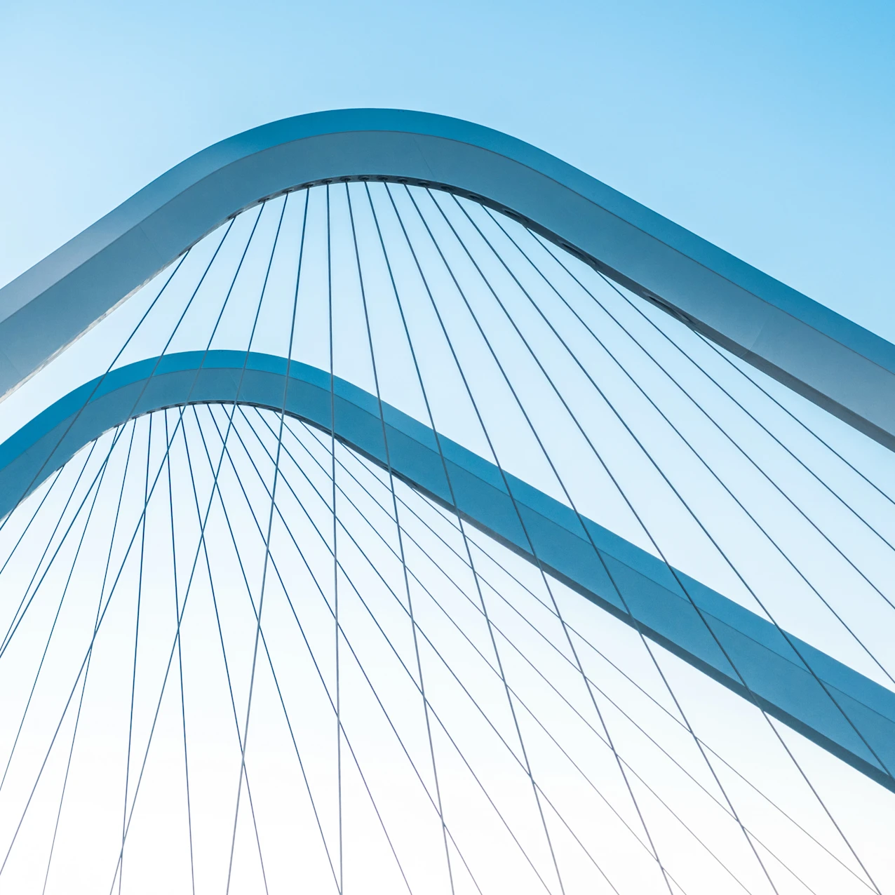 Close-up of two curved white metal arches with parallel cables against a clear blue sky, forming a modern bridge design.