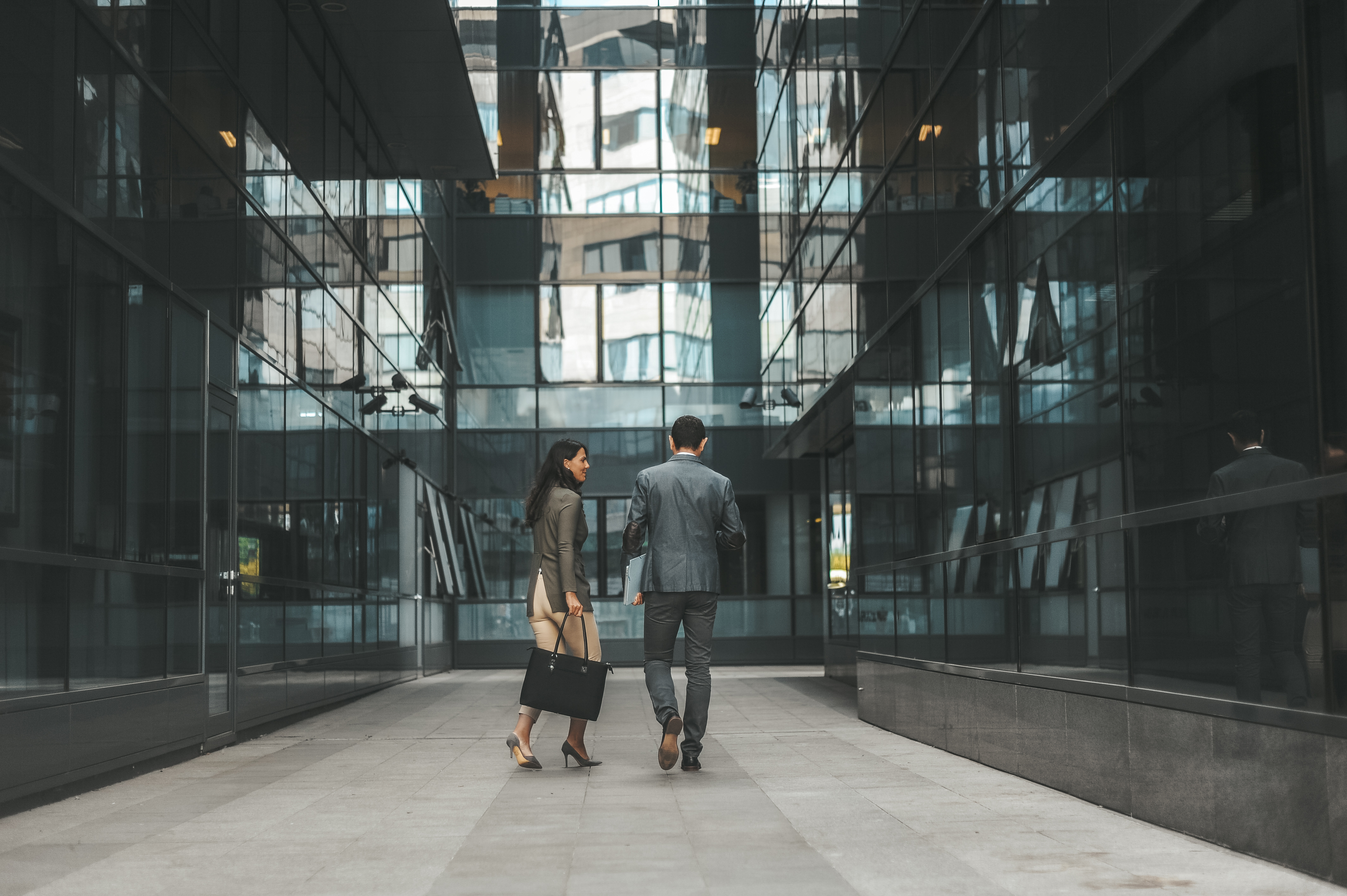 A man and a woman in business attire walking between modern glass buildings in an urban setting, seen from behind.
