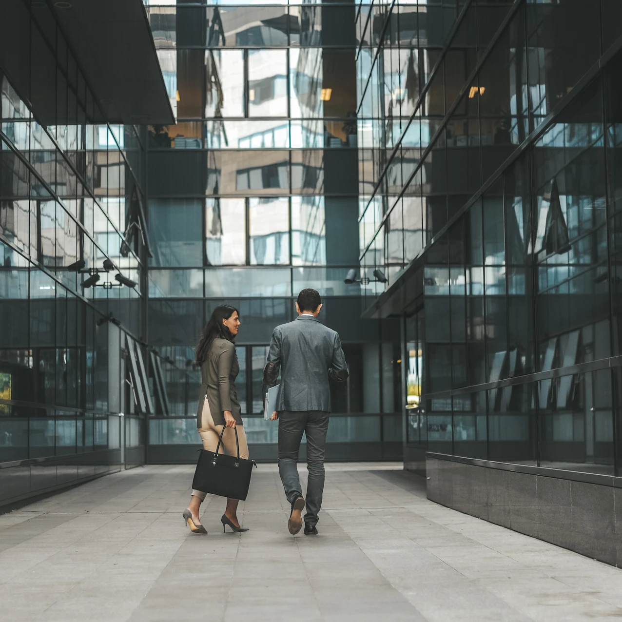 A man and a woman in business attire walking between modern glass buildings in an urban setting, seen from behind.