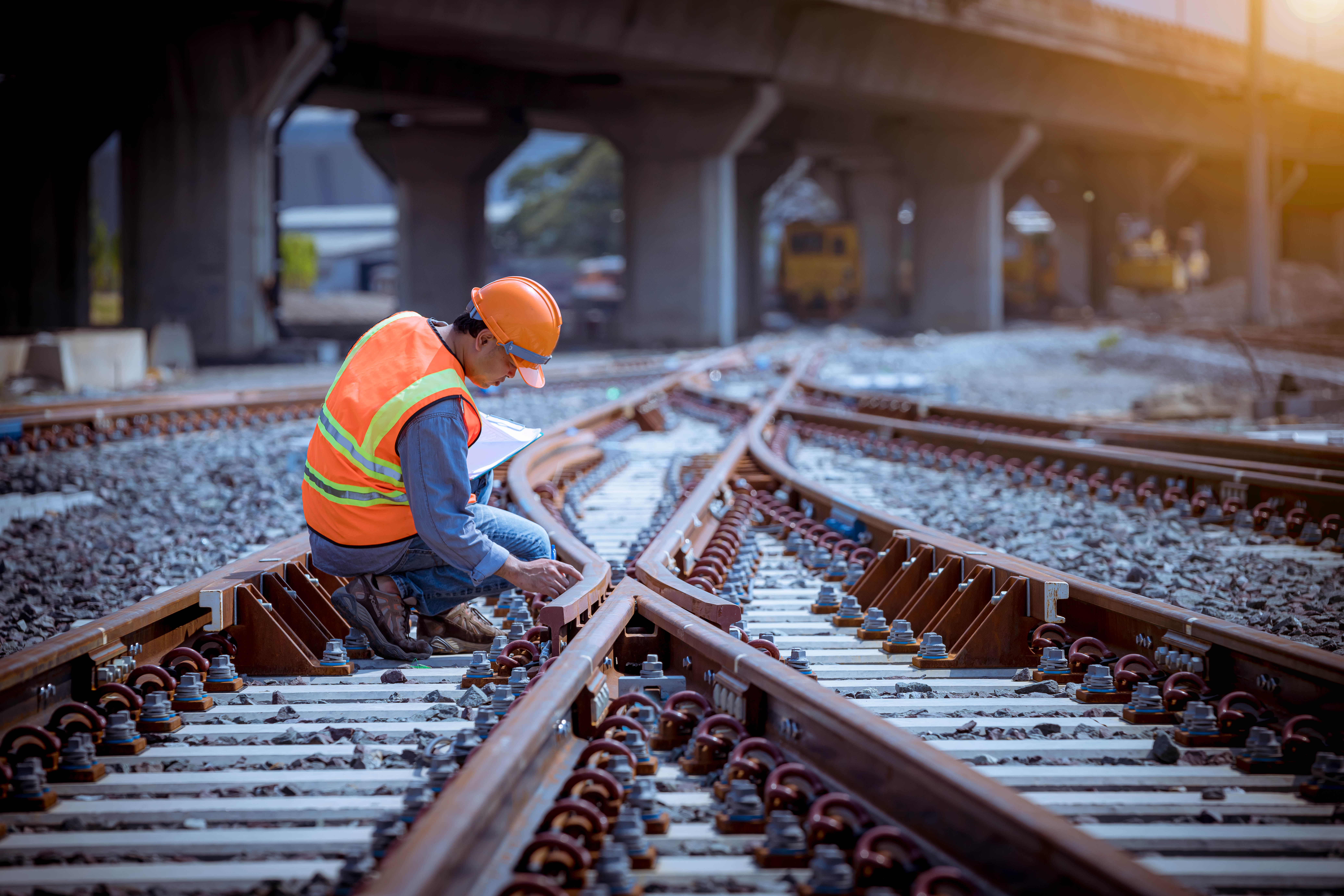 A rail worker wearing an orange safety vest and helmet inspects railroad tracks under a bridge during daylight.