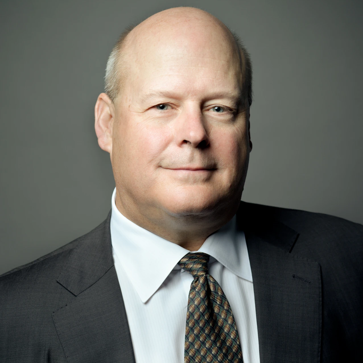 David Beckman wearing a dark suit jacket, white dress shirt, and patterned tie standing against a plain gray background.