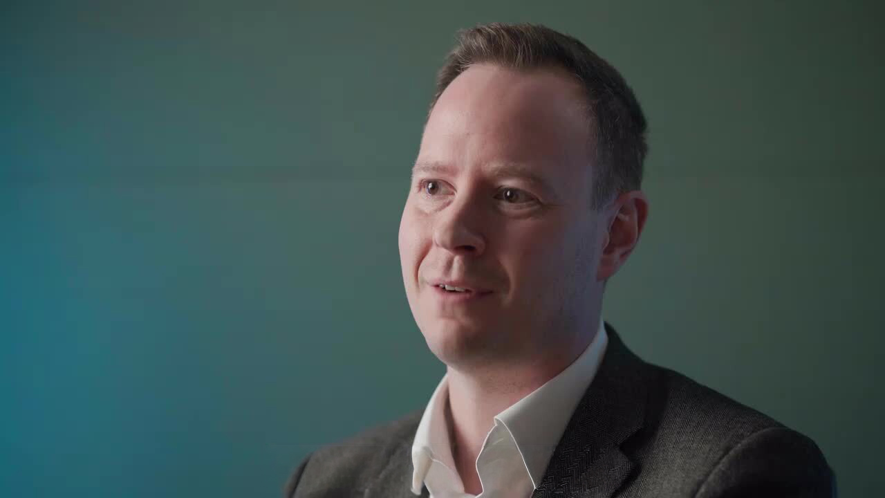 Jon Priestley speaking during a video interview, wearing a white shirt and grey blazer against a teal background.