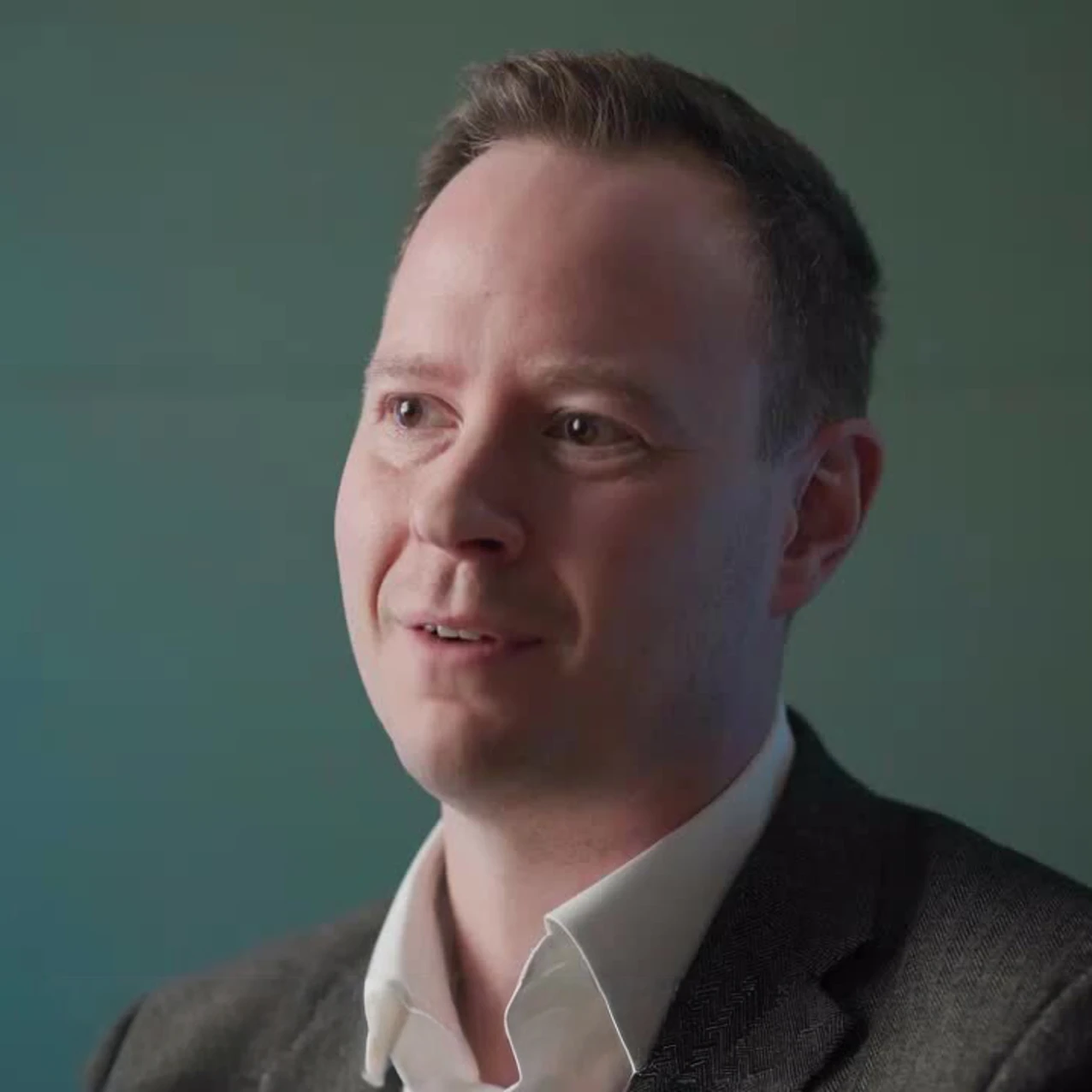 Jon Priestley speaking during a video interview, wearing a white shirt and grey blazer against a teal background.