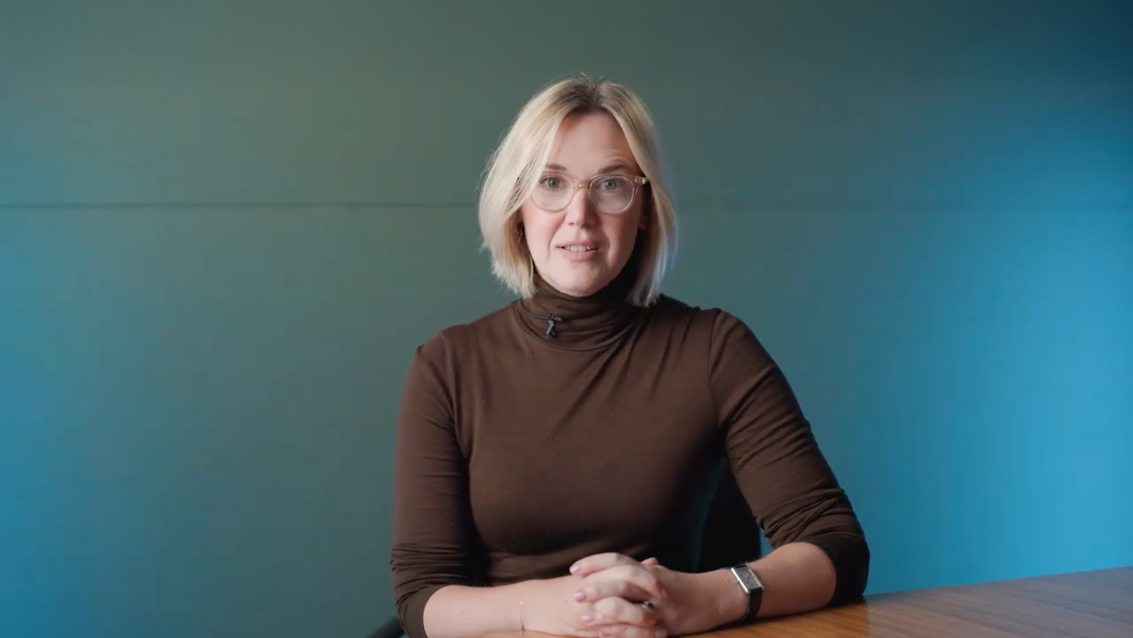 Person with blonde hair wearing a brown turtleneck sits at a wooden table with hands clasped, against a teal background.