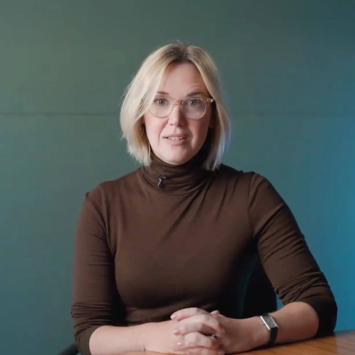 Person with blonde hair wearing a brown turtleneck sits at a wooden table with hands clasped, against a teal background.