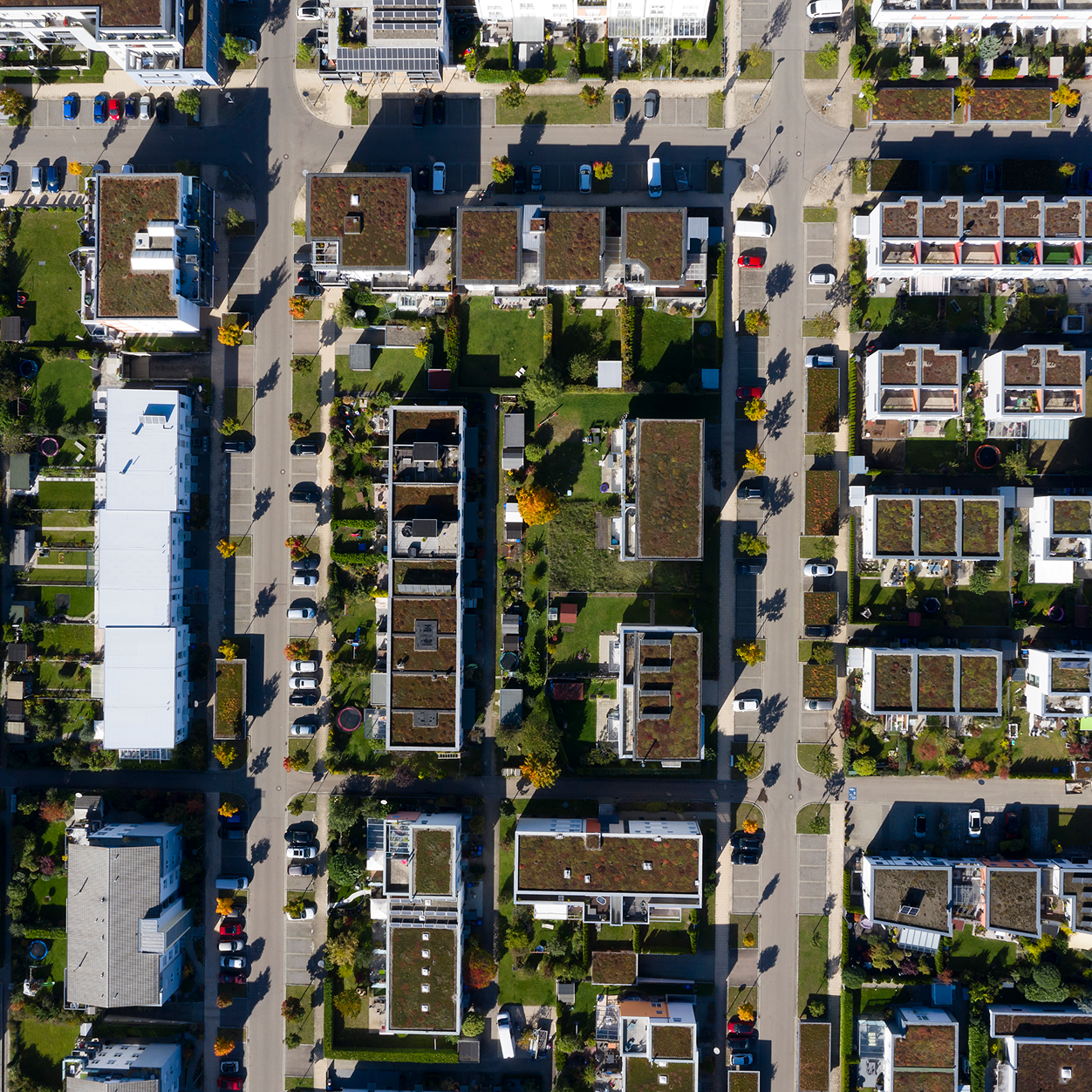 Aerial view of a residential neighborhood with rows of modern houses featuring green roofs, gardens, parked cars, and tree-lined streets on a sunny day.