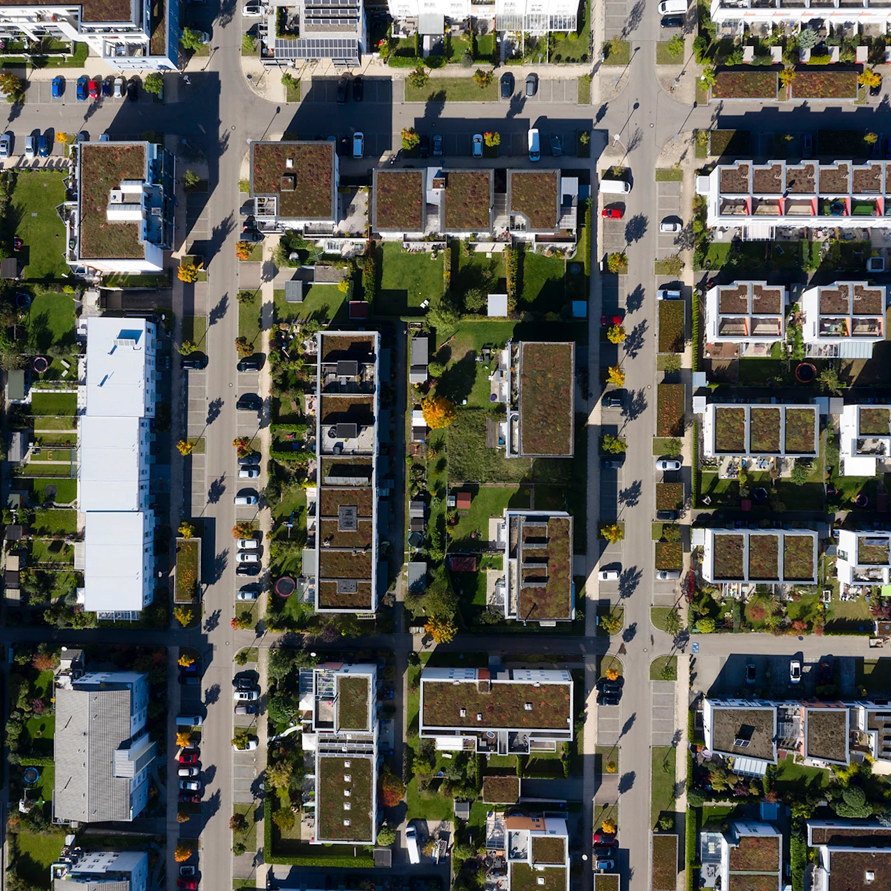 Aerial view of a residential neighborhood with rows of modern houses featuring green roofs, gardens, parked cars, and tree-lined streets on a sunny day.