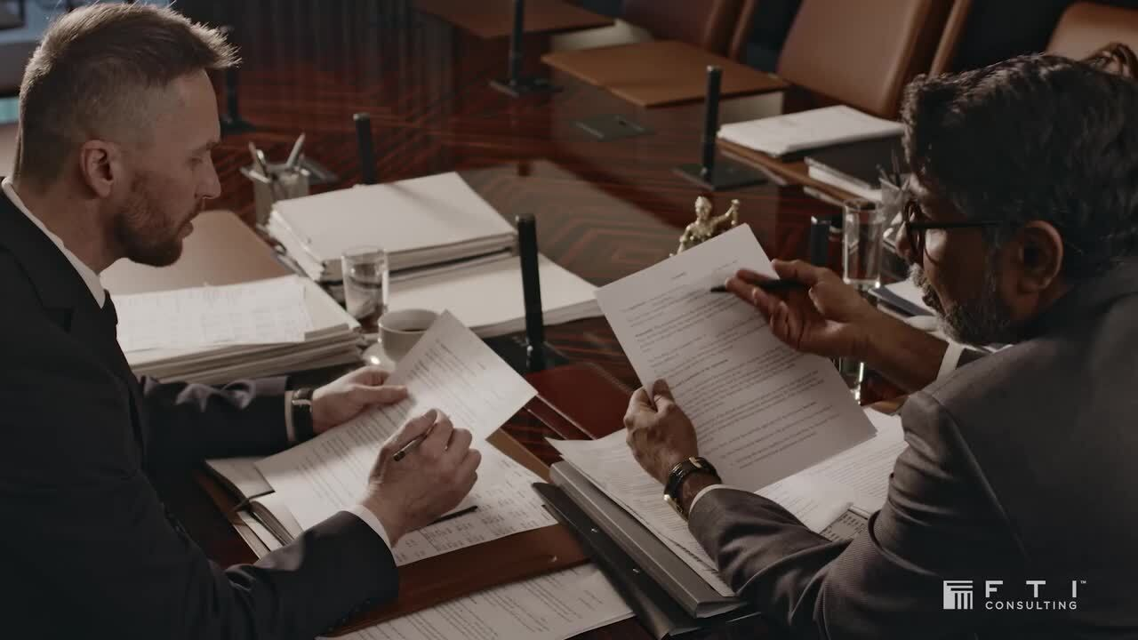 Two men in suits reviewing and marking documents over a dark wooden conference table filled with papers, binders, glasses of water, and a coffee cup.
