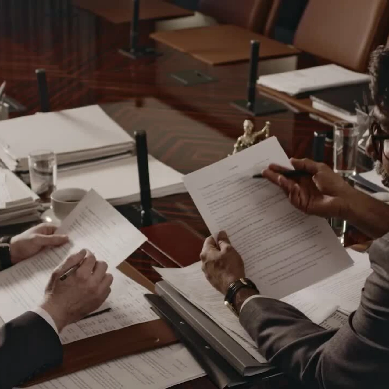 Two men in suits reviewing and marking documents over a dark wooden conference table filled with papers, binders, glasses of water, and a coffee cup.