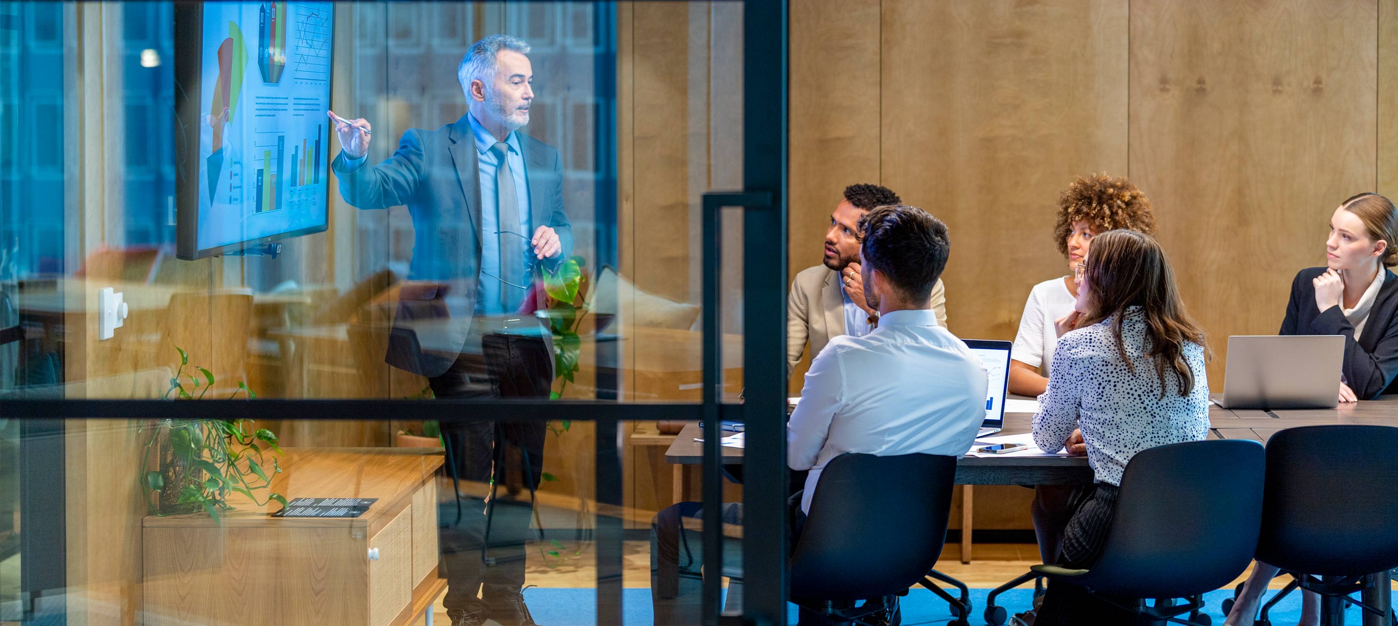 A man presents charts on a screen to a seated group in a glass-walled meeting room, with laptops on the table.