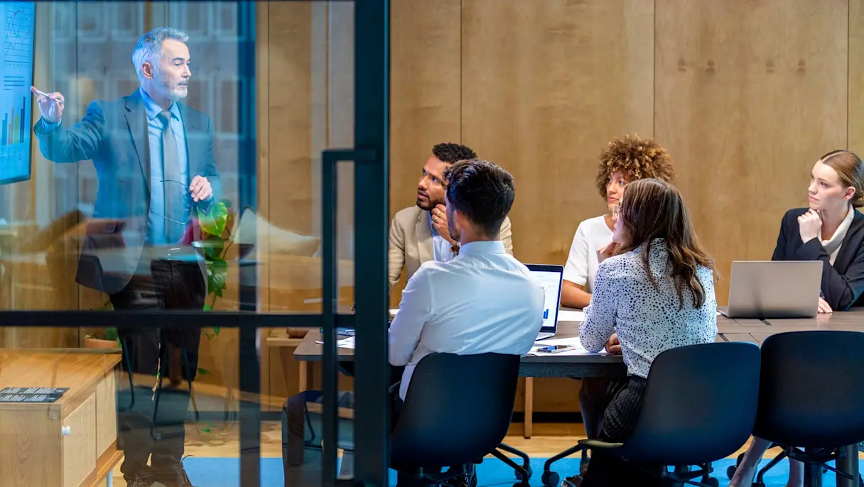 A man presents charts on a screen to a seated group in a glass-walled meeting room, with laptops on the table.