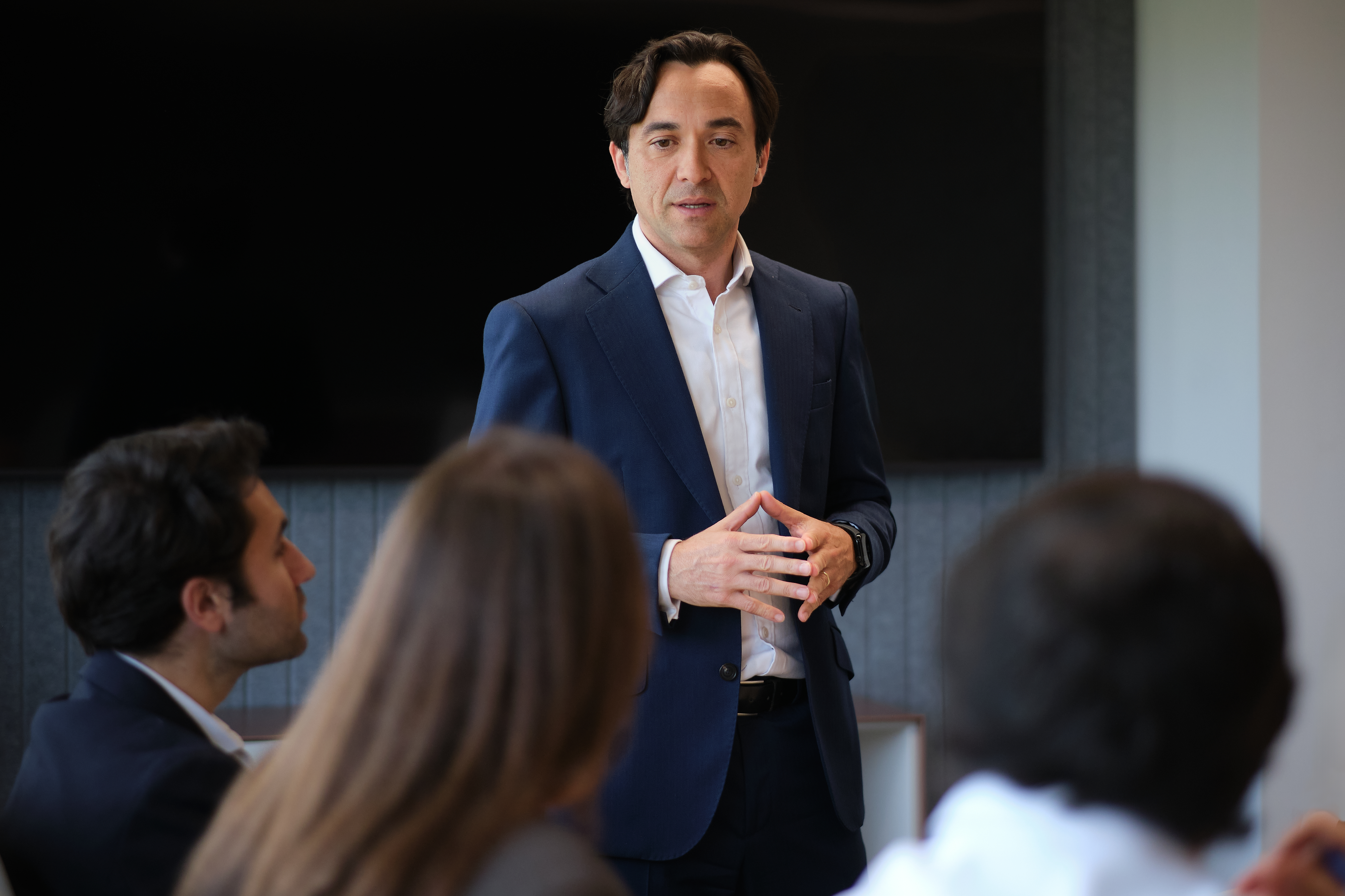 A man in a navy suit and white shirt stands and speaks to three seated people in a modern office meeting room with a large black screen on the wall.