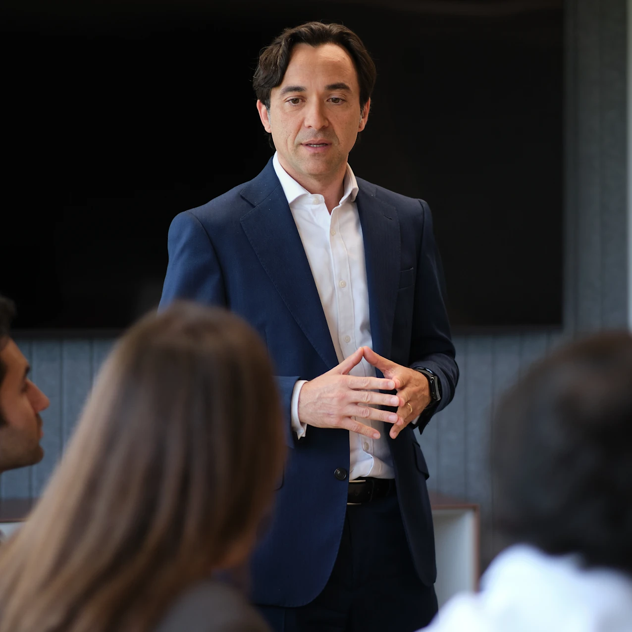 A man in a navy suit and white shirt stands and speaks to three seated people in a modern office meeting room with a large black screen on the wall.