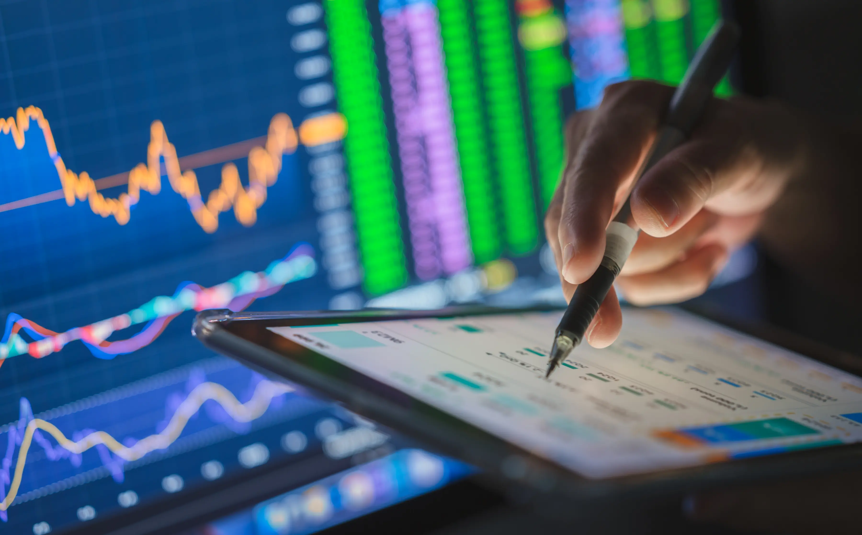 A hand holding a pen interacts with a tablet displaying financial data, with colorful stock market charts and graphs visible on a screen in the background.