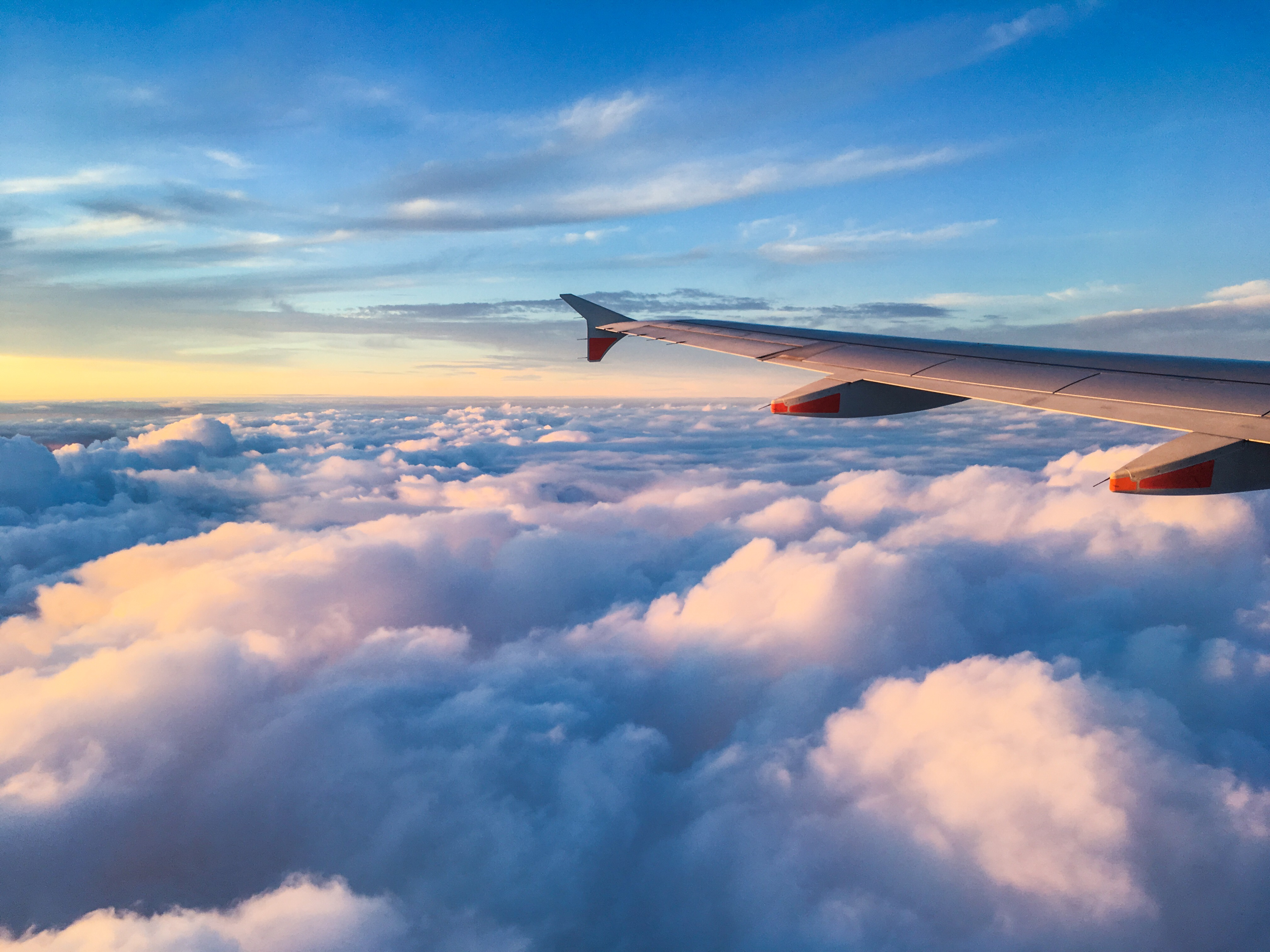 View from airplane window showing wing above fluffy clouds illuminated by sunset under a blue sky.