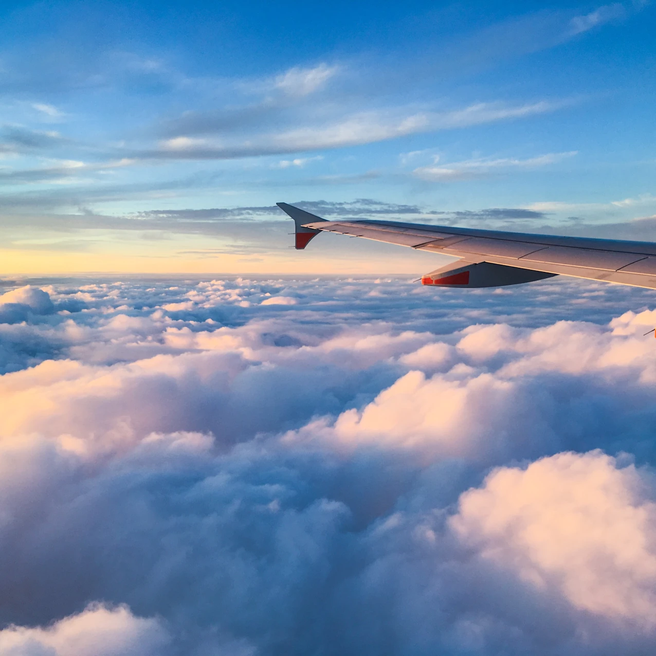 View from airplane window showing wing above fluffy clouds illuminated by sunset under a blue sky.