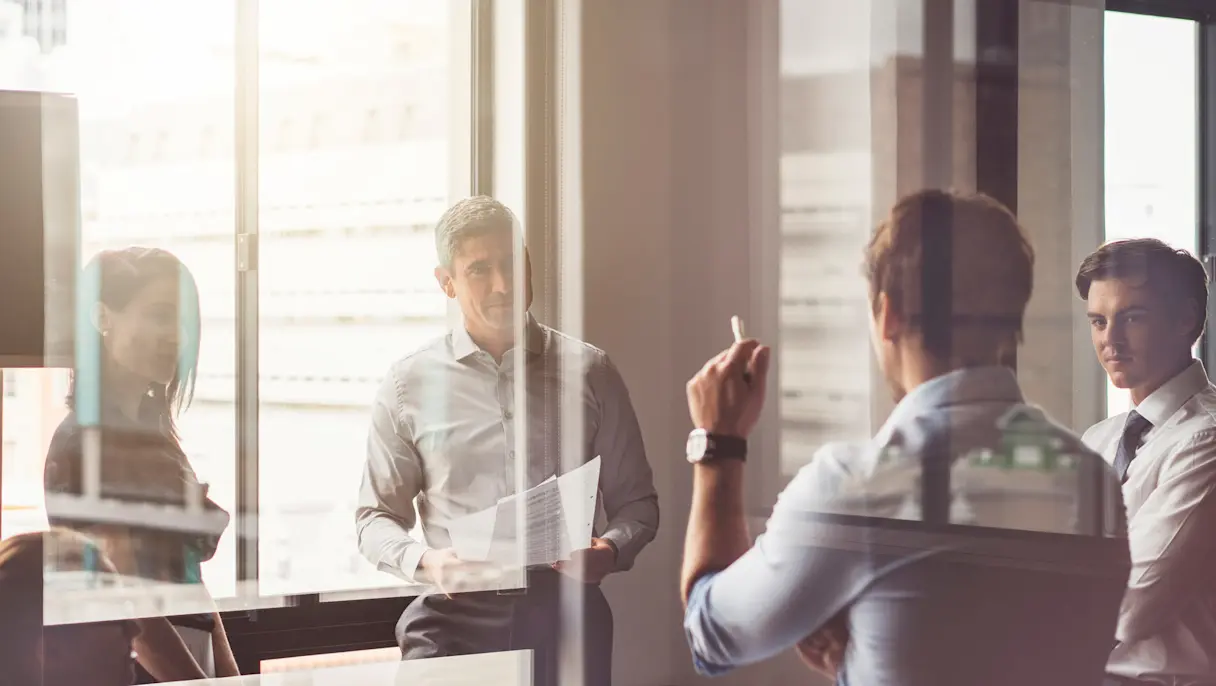 A group of four professionals standing in a glass-walled office, engaged in a discussion, with one person holding a document while another gestures as if explaining something.