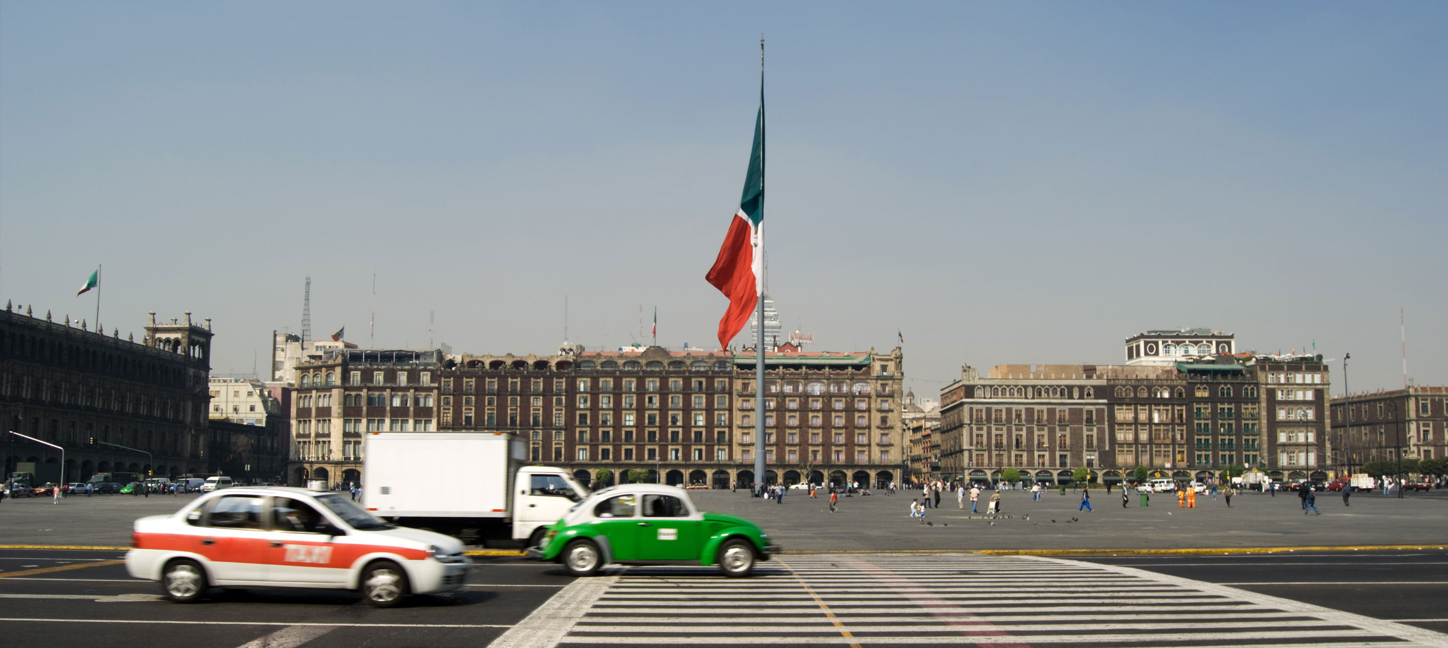 Cars and taxis cross a zebra crossing in Mexico City’s Zócalo with a large Mexican flag and historic buildings.