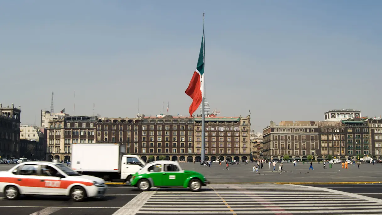 Cars and taxis cross a zebra crossing in Mexico City’s Zócalo with a large Mexican flag and historic buildings.