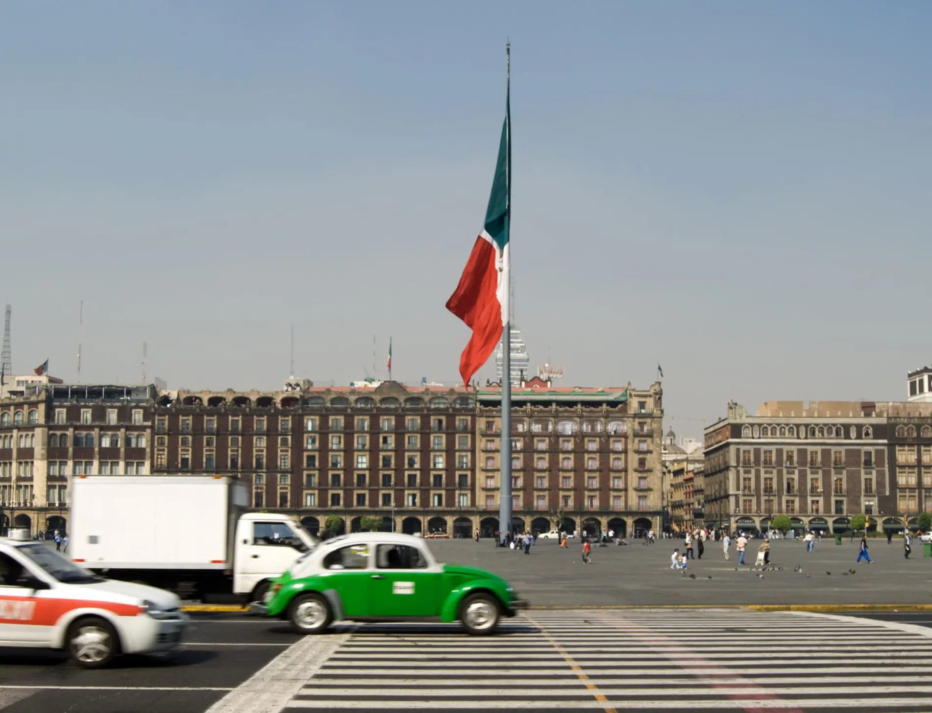 Cars and taxis cross a zebra crossing in Mexico City’s Zócalo with a large Mexican flag and historic buildings.