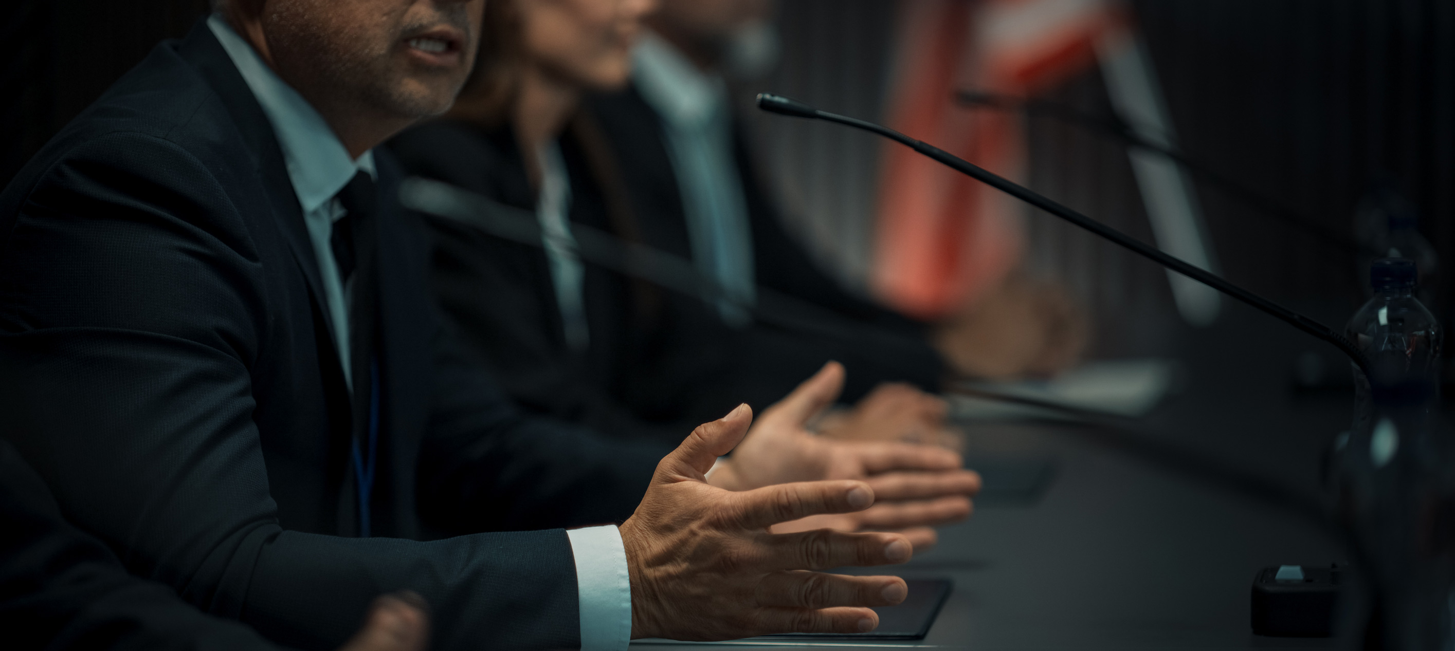 Close-up of panellists in suits at a table, one gesturing while speaking into a microphone, red banner blurred behind.