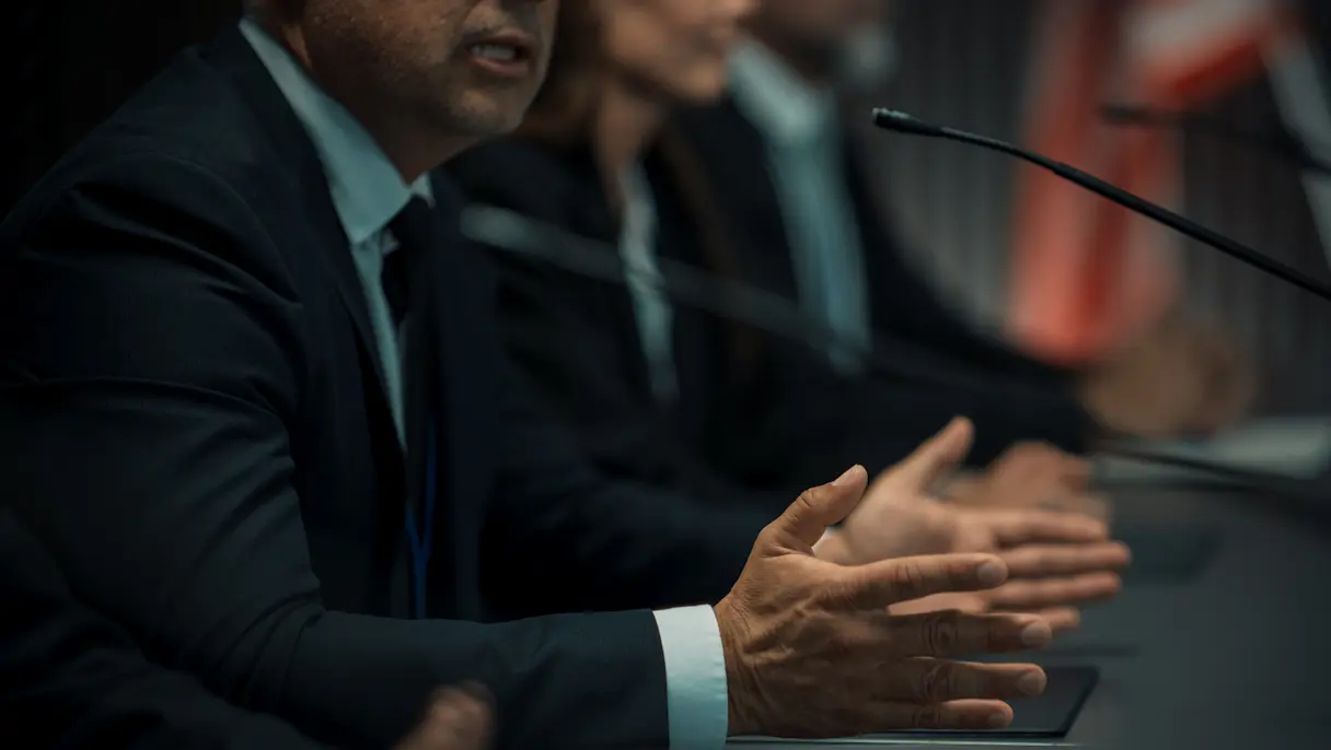 Close-up of panellists in suits at a table, one gesturing while speaking into a microphone, red banner blurred behind.