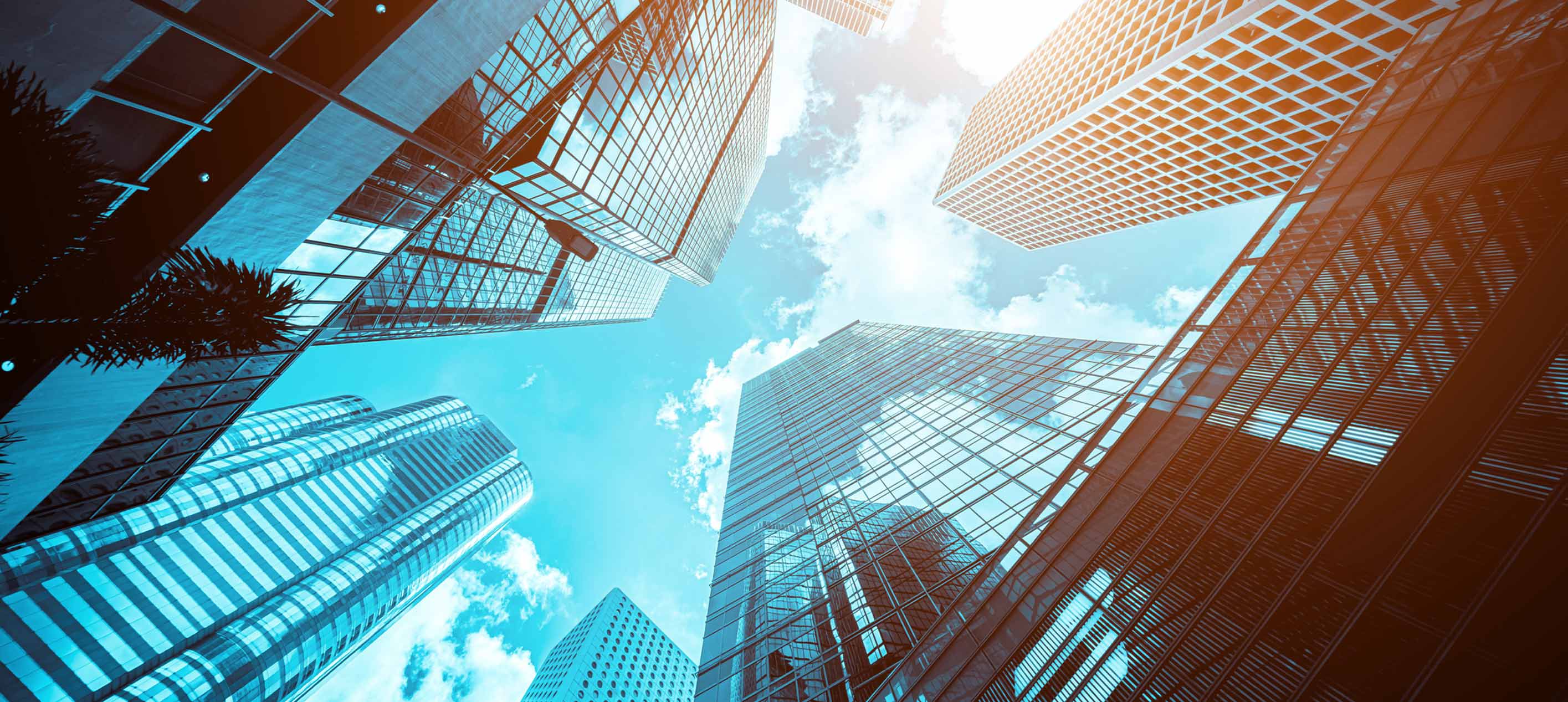 Upward view of glass skyscrapers against a blue sky with clouds, in blue and orange toned photography.