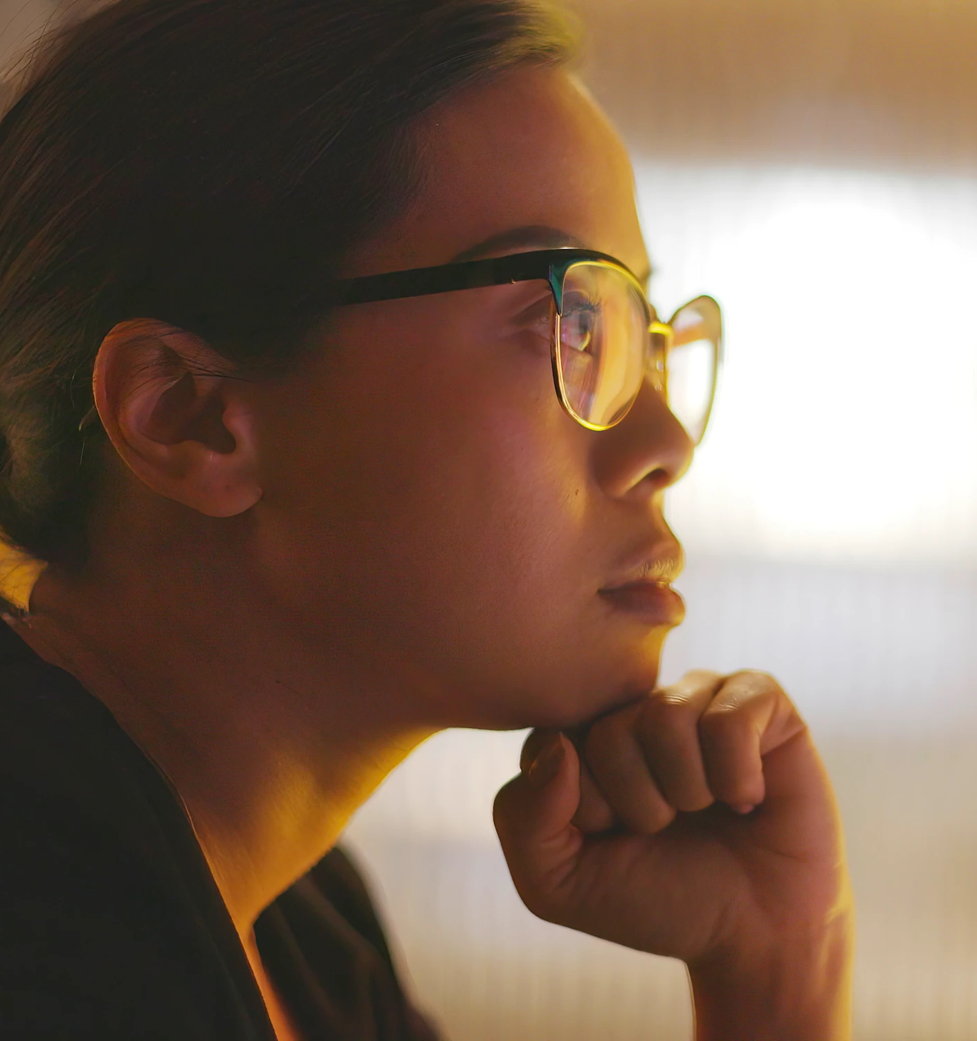 Close-up side profile of a person wearing glasses, resting their chin on their fist, looking thoughtfully into the light source.
